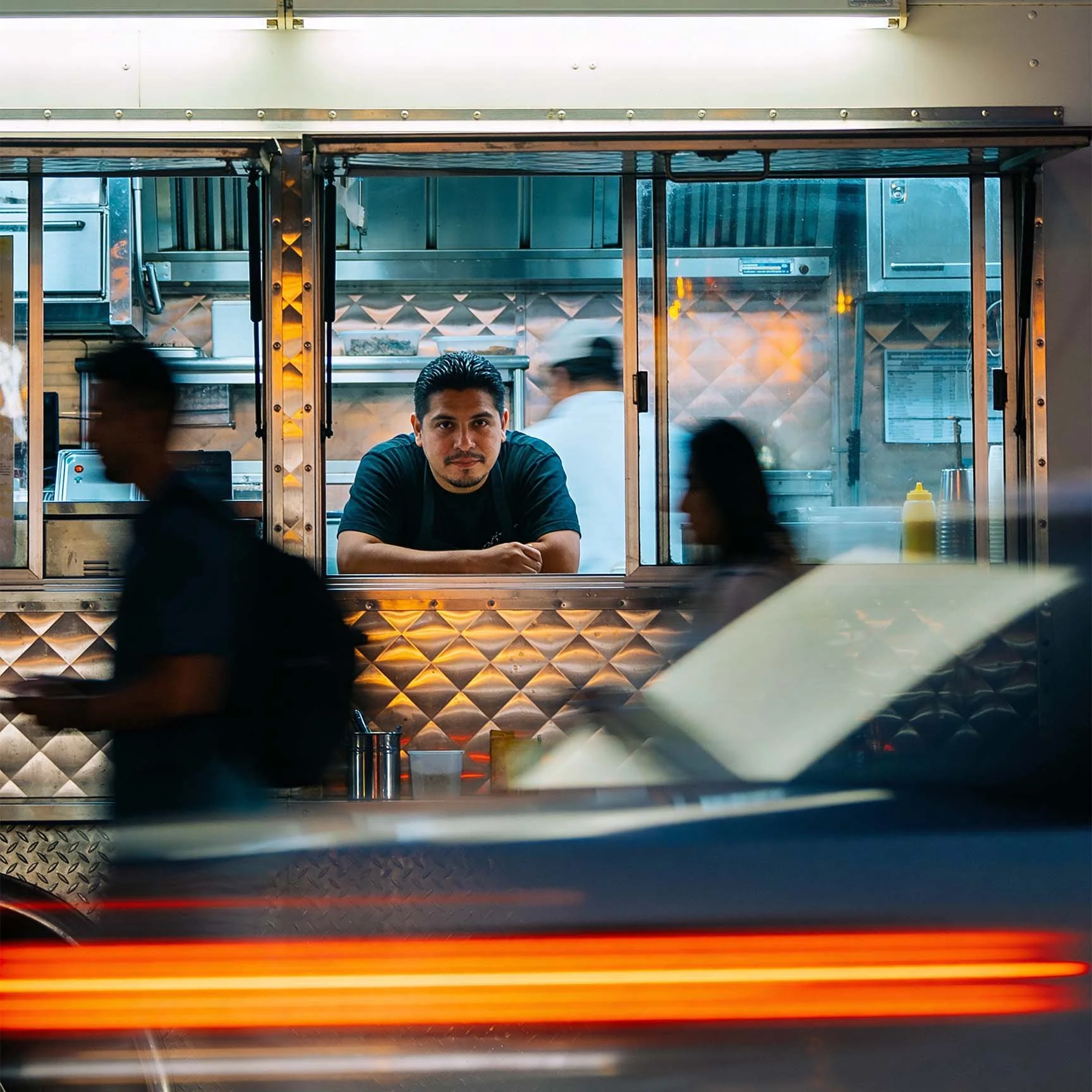 A man leaning on a food truck counter, looking out through the window at passing blurred figures and a vehicle with streaks of red light.
