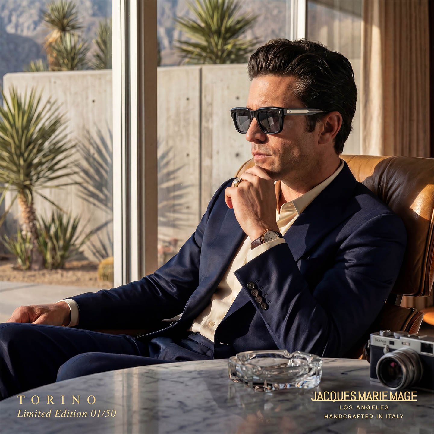 A man in a navy suit and sunglasses sitting in a sunlit room by large windows with desert plants outside. He has dark hair, a watch, a ring, and a contemplative expression, with a camera and ashtray on the table in front of him.