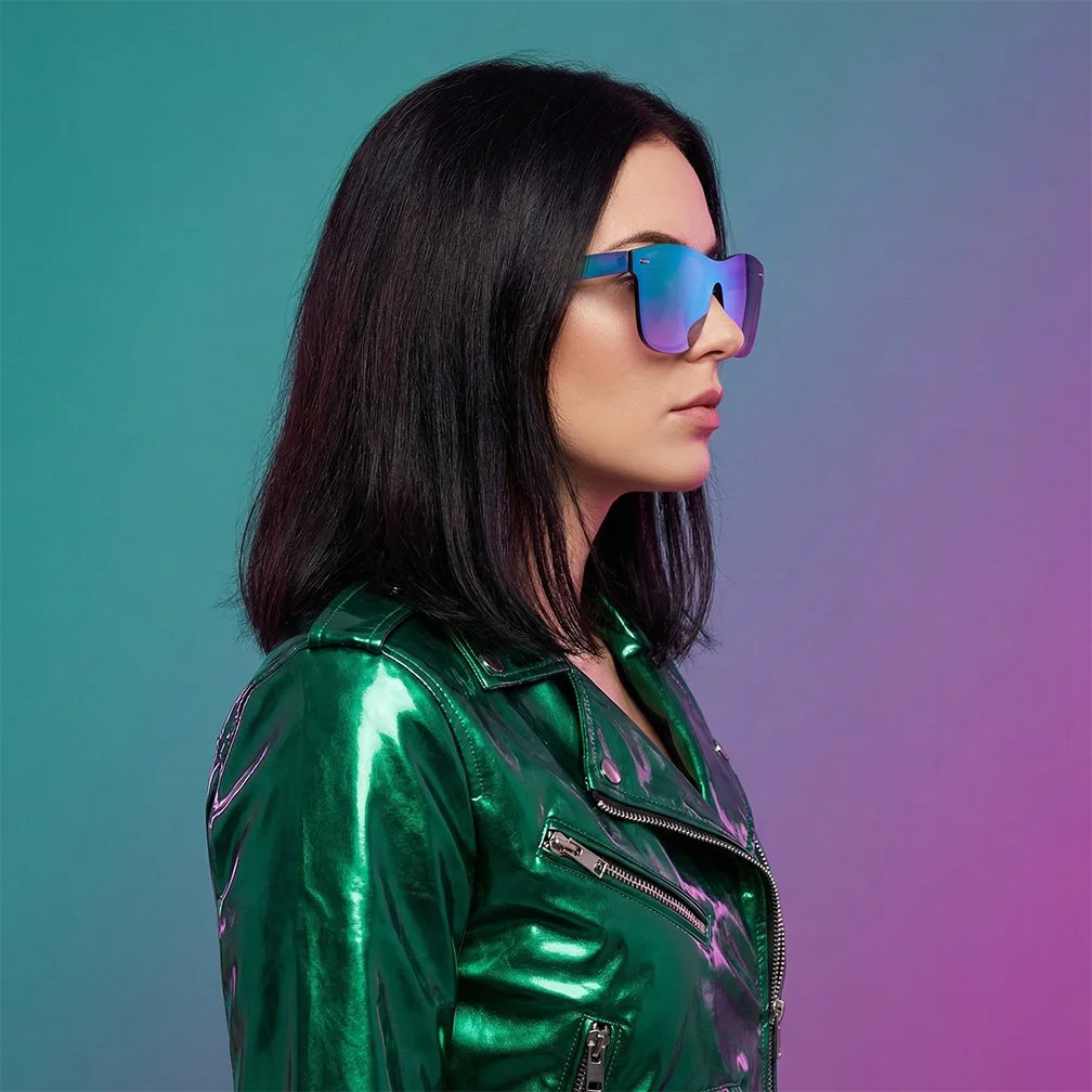 A woman with shoulder-length dark hair wearing mirrored sunglasses and a shiny green leather jacket, posing in front of a gradient background.