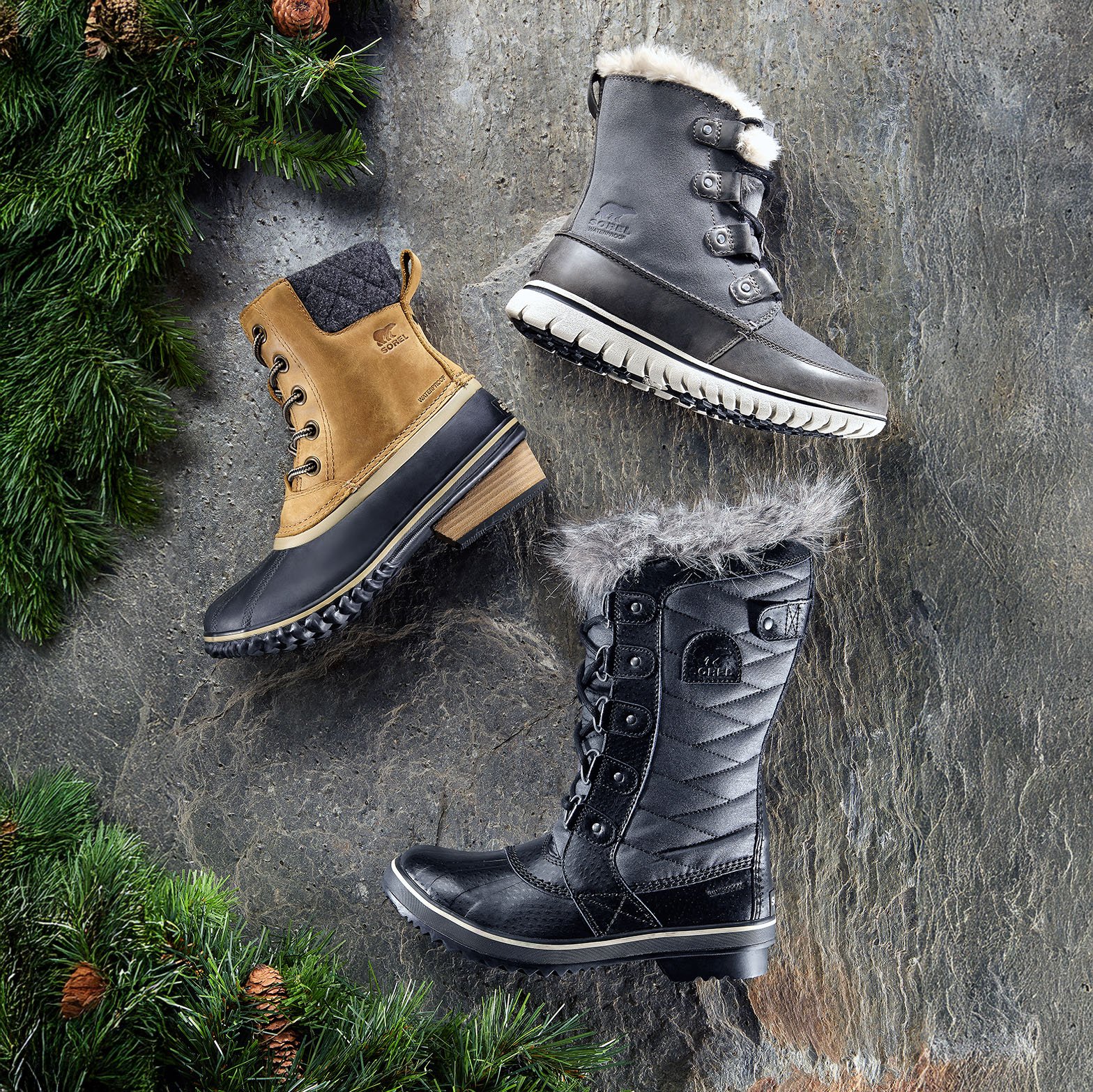Three pairs of winter boots arranged on a stone surface with green pine branches nearby. One is a light brown, waterproof style with a black sole, one is a gray, insulated style with a white fluffy lining, and the third is a black, quilted waterproof