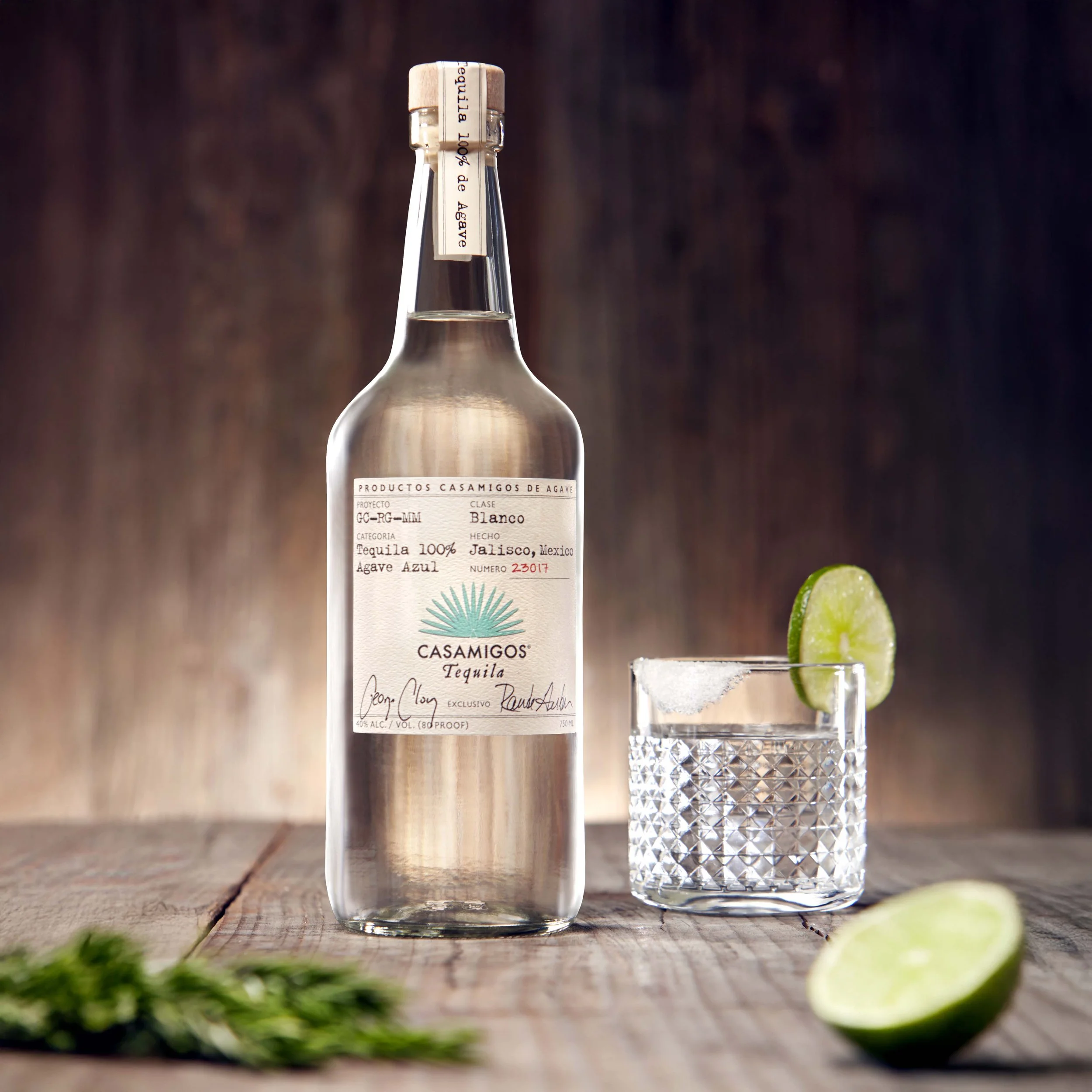 A glass of tequila with ice and a lime wedge on top, placed next to a bottle of Casamigos tequila on a wooden surface.