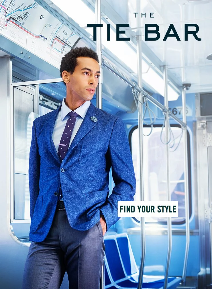 A man in a blue blazer and navy pants standing inside a train, looking out the window. Text on the image says 'The Tie Bar' and 'Find Your Style'.