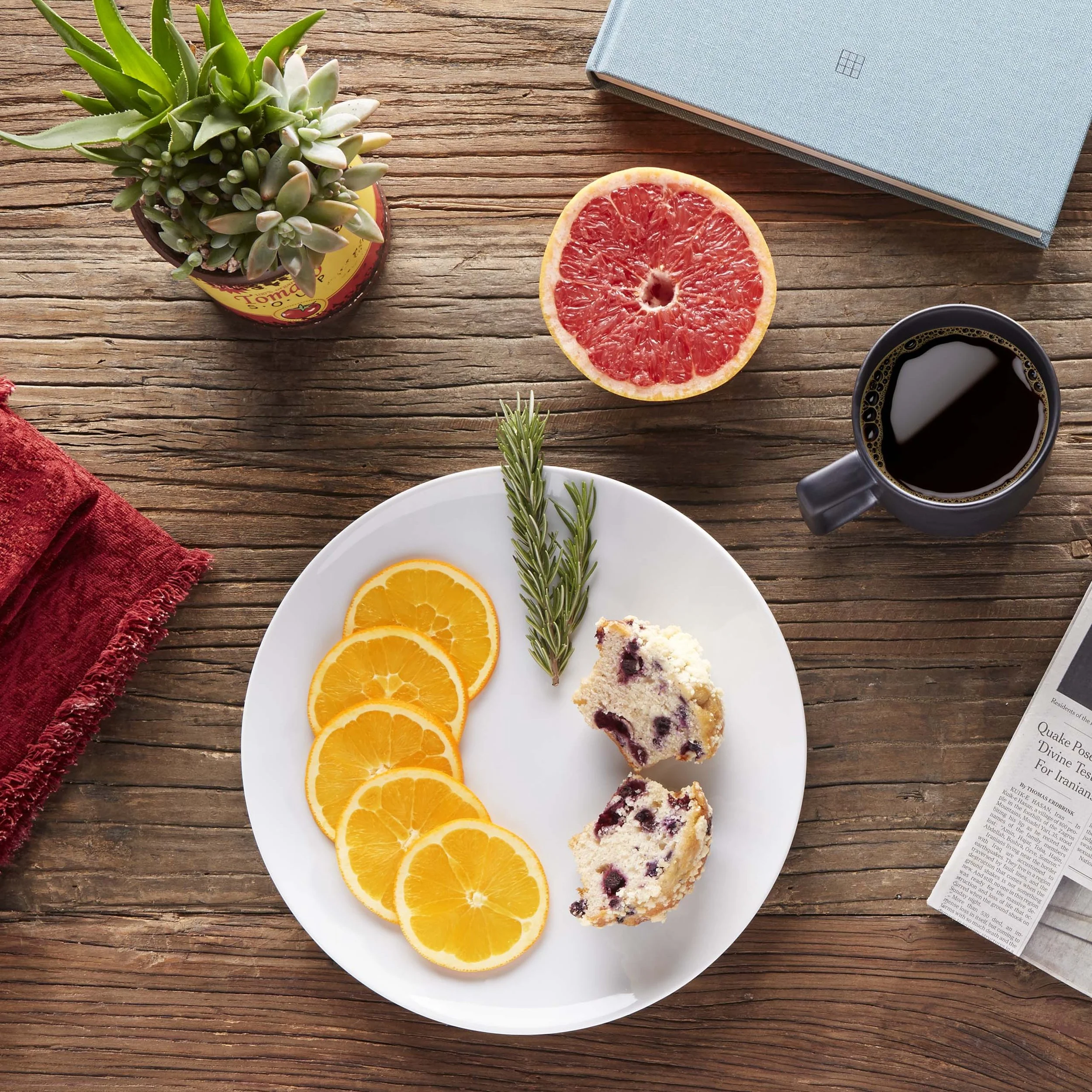A top-down view of a breakfast table with a white plate containing orange slices, a sprig of rosemary, and blueberry scone halves. Surrounding the plate are a grapefruit half, black coffee in a mug, a potted succulent, a closed blue notebook, a red c