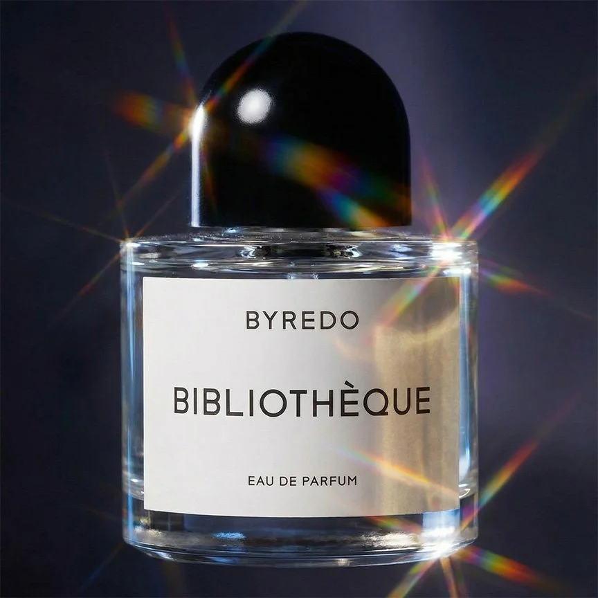 A clear glass bottle of Byredo Bibliothèque Eau de Parfum with a black cap, displaying a white label with black text, reflecting light with colorful prismatic flares.