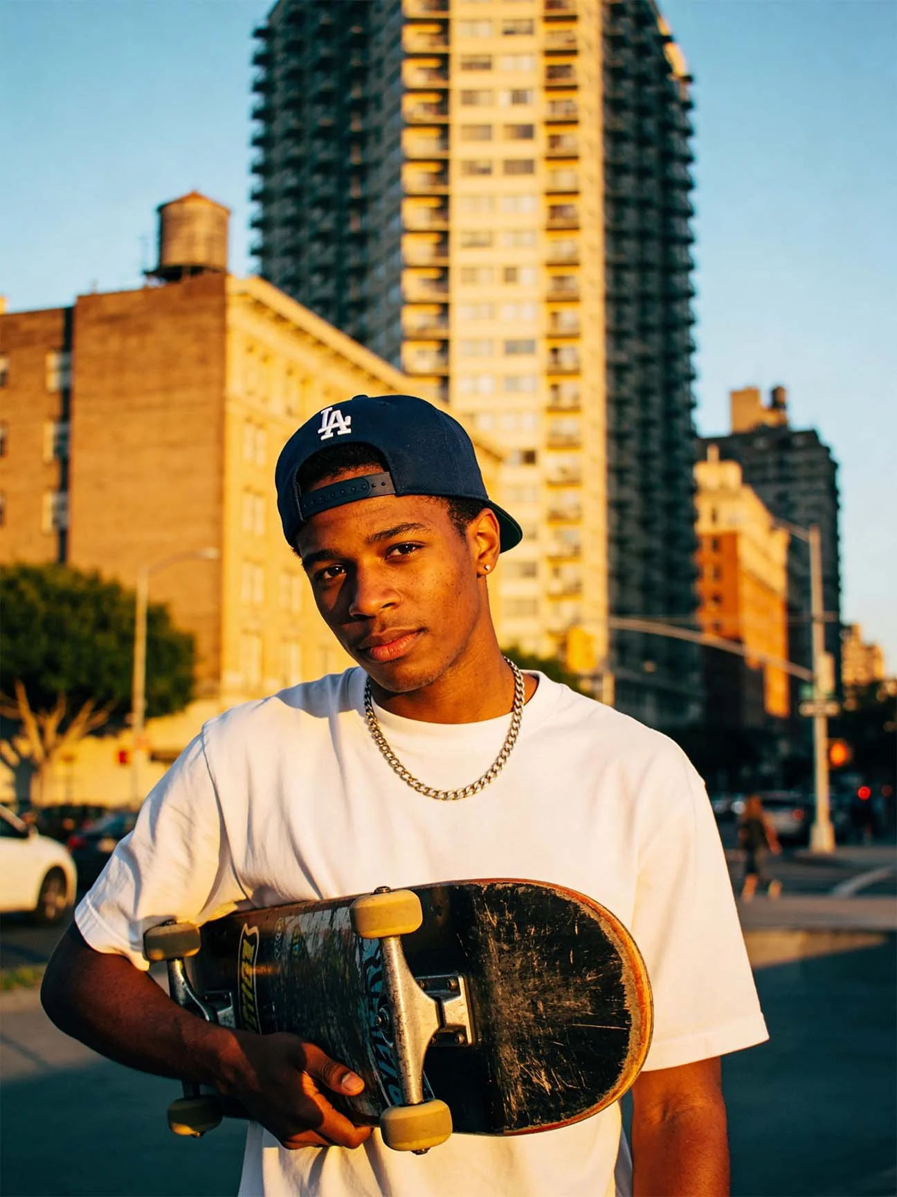 A young man wearing a white T-shirt, a navy blue baseball cap turned backward, and a silver chain necklace, holding a skateboard with yellow wheels, standing in an urban city environment during sunset.