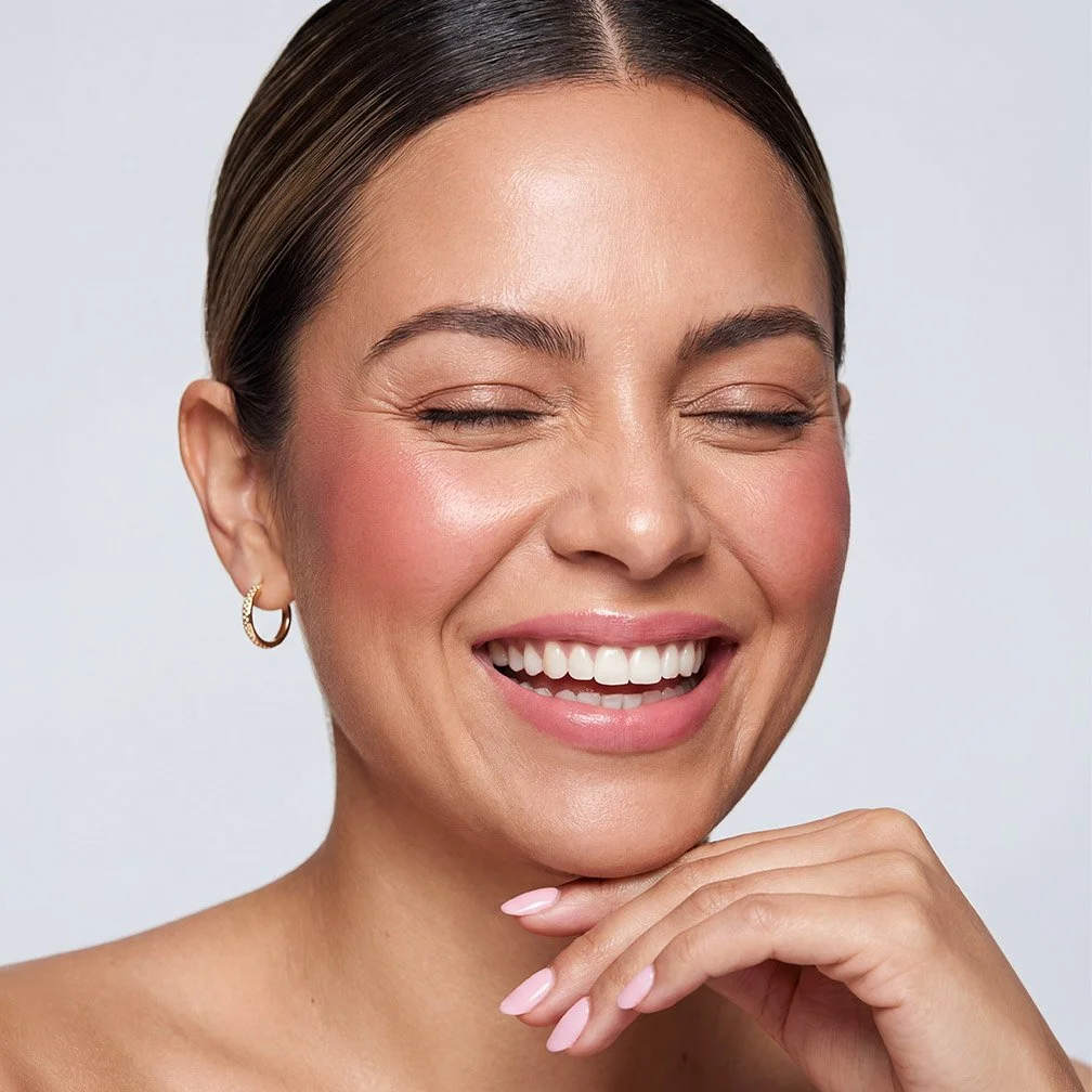 Close-up of a smiling woman with smooth skin, her eyes closed, wearing a small earring, and her hand gently touching her chin against a plain background.