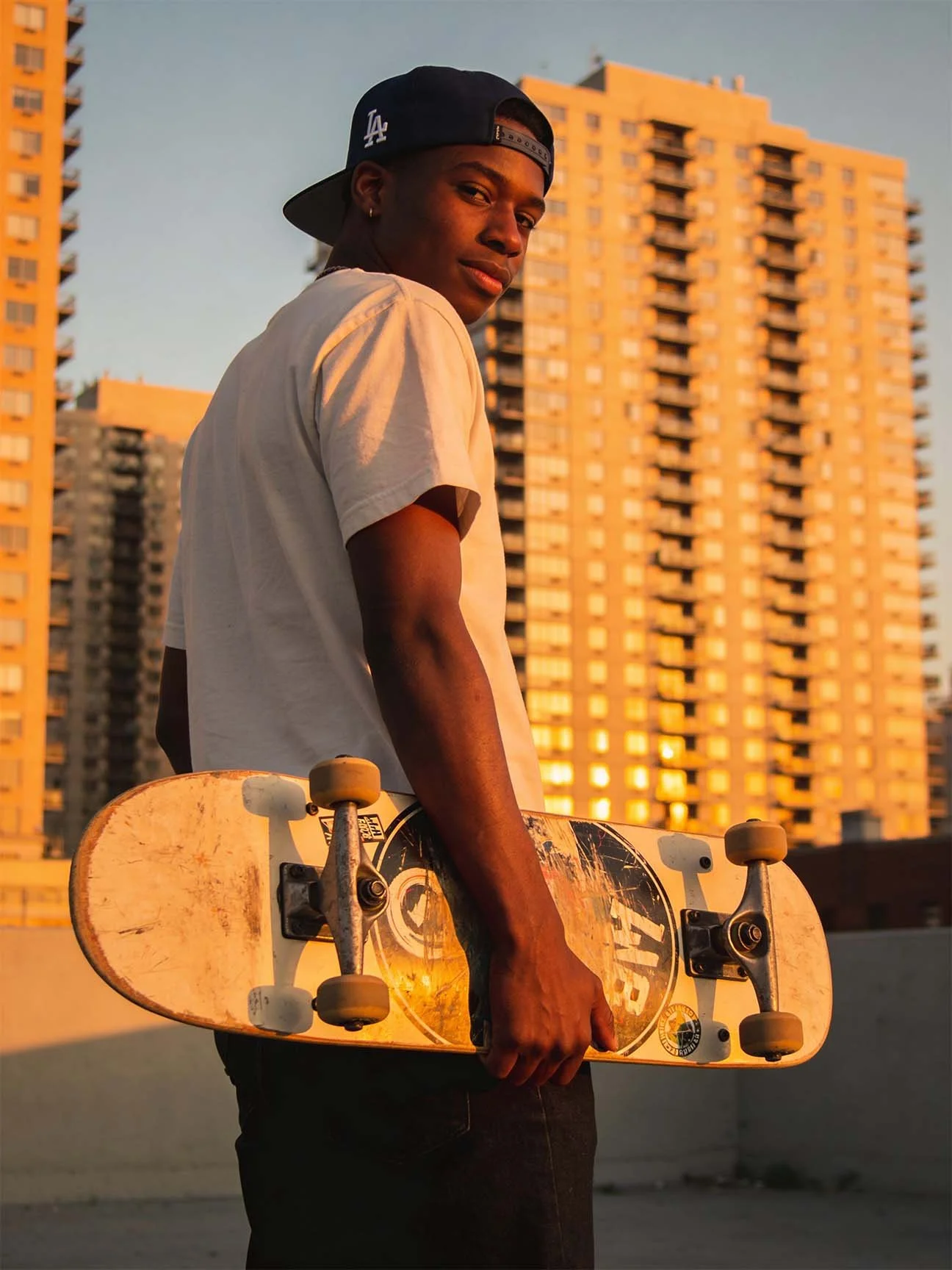 A young man wearing a navy blue cap with LA logo and a beige t-shirt holding a skateboard on a rooftop at sunset with tall apartment buildings in the background.