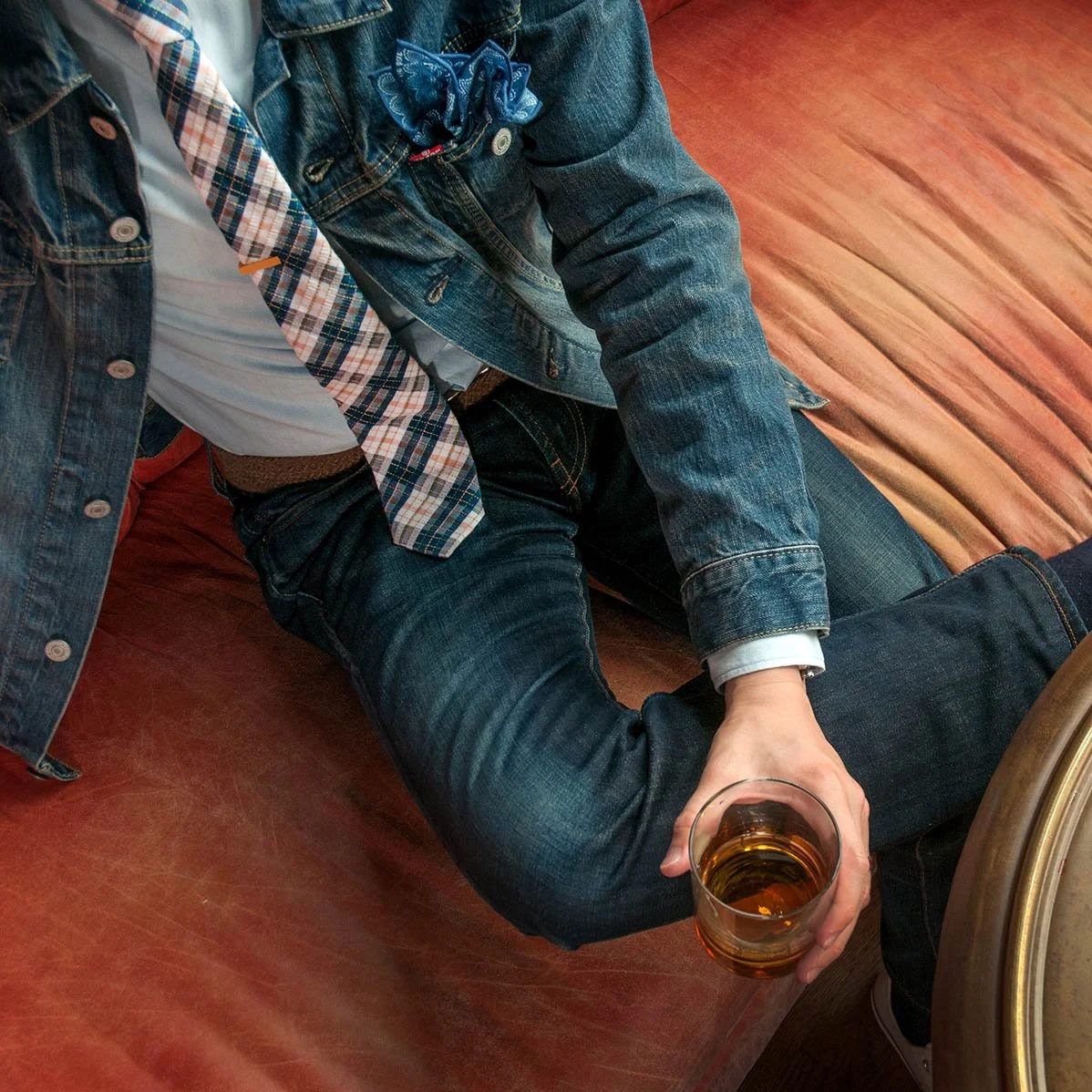 Person sitting on a wooden floor, wearing denim jacket, plaid tie, and dark jeans, holding a glass of beer.