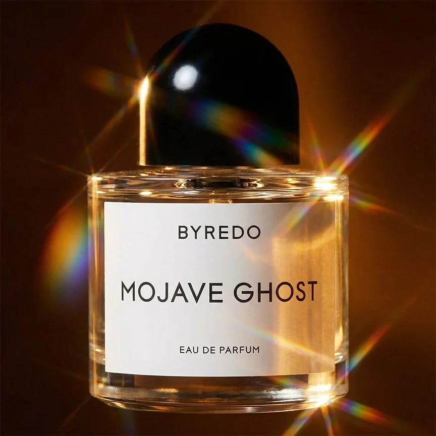 A clear glass perfume bottle labeled BYREDO MOJAVE GHOST with a black cap, illuminated by light creating rainbow-colored lens flares.