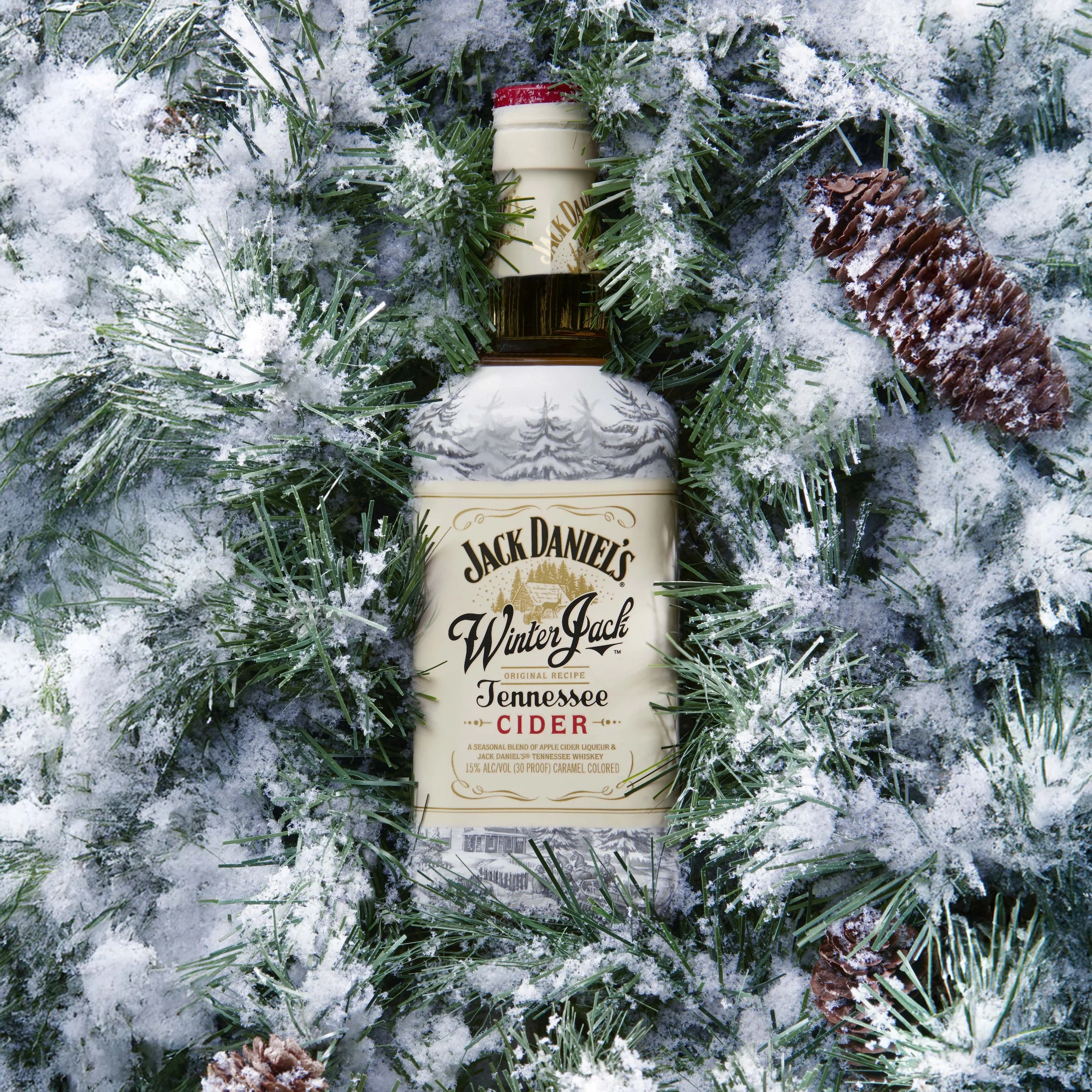 A bottle of Jack Daniel's Winter Jack Tennessee Cider surrounded by snow-covered pine branches and pinecones.