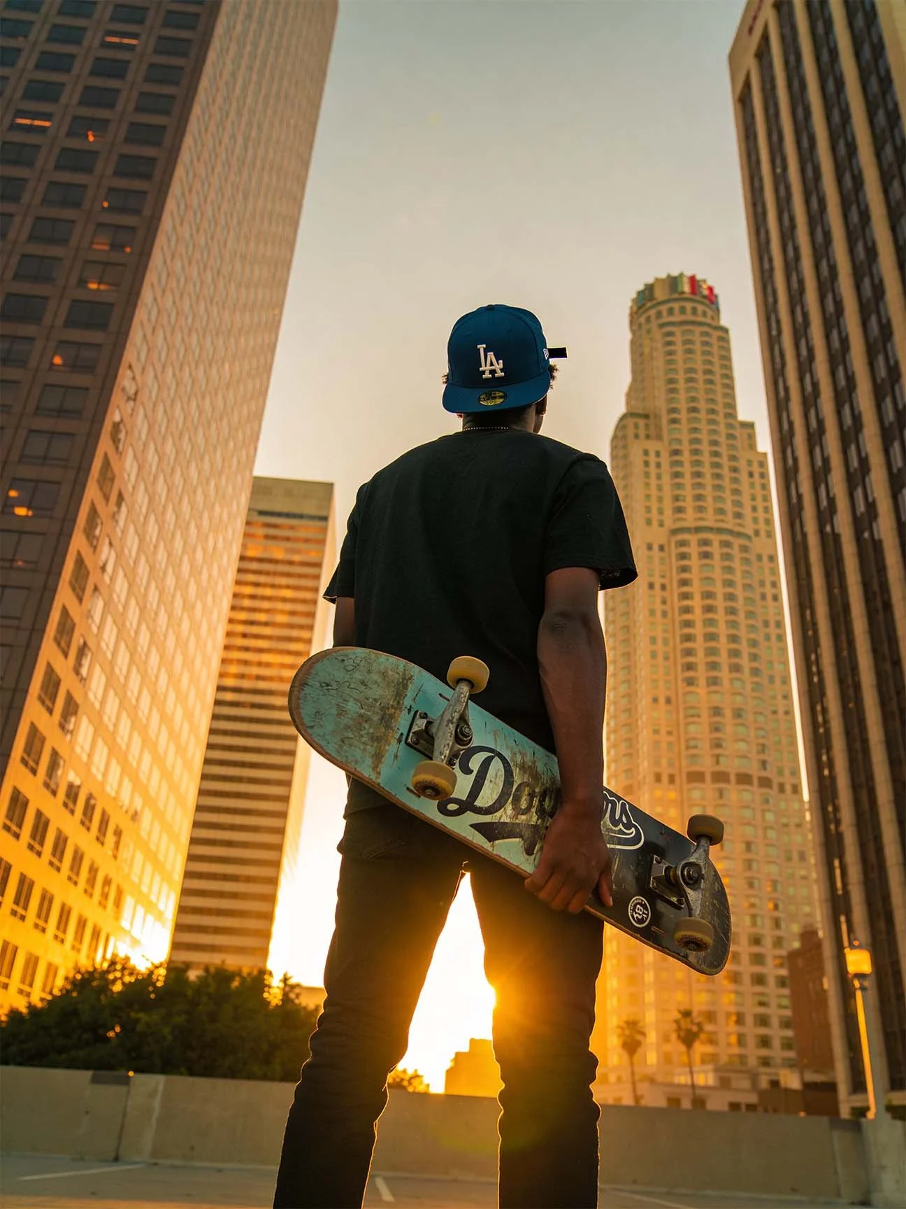 A young man wearing a blue LA baseball cap, black t-shirt, and jeans stands holding a skateboard, facing tall buildings during sunset in an urban setting.