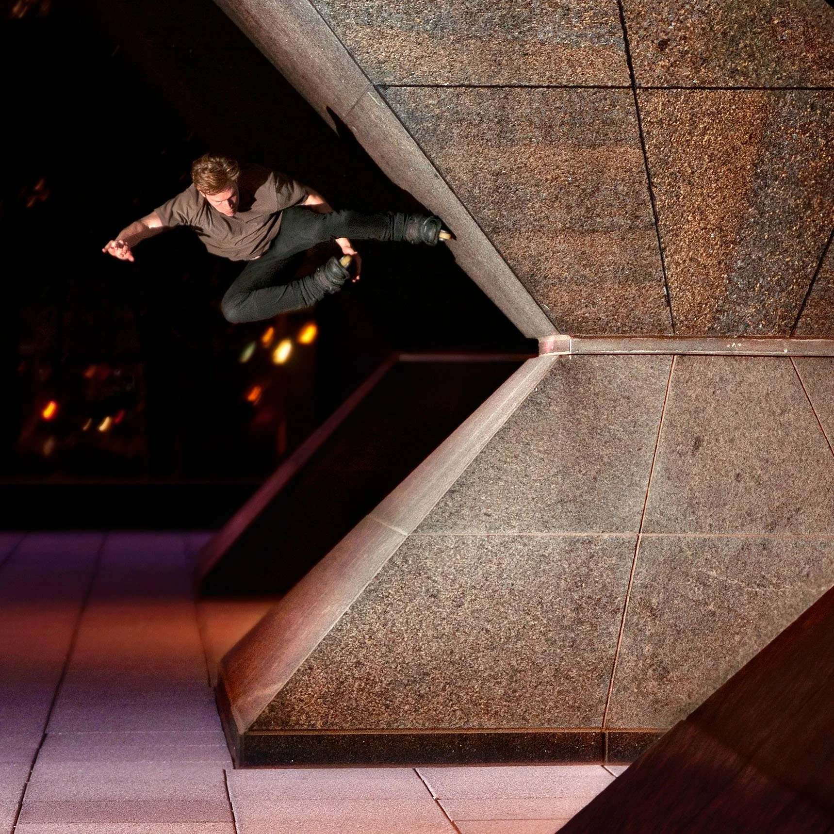 A man is mid-air in a jump on a rooftop or ledge at night, with city lights in the background.