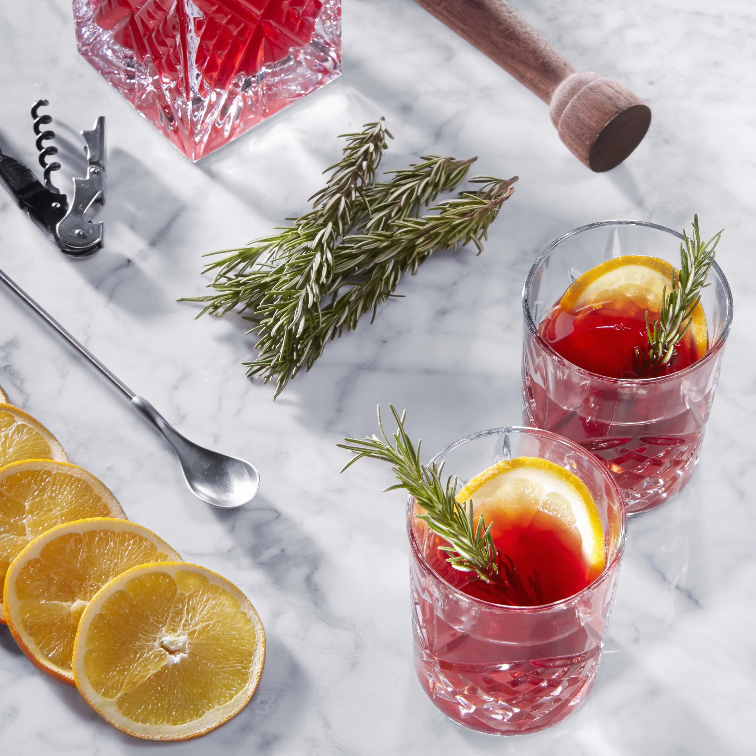 Two glasses of red beverage garnished with orange slices and rosemary sprigs on a marble surface, with sliced oranges, a corkscrew, and sprigs of rosemary nearby.
