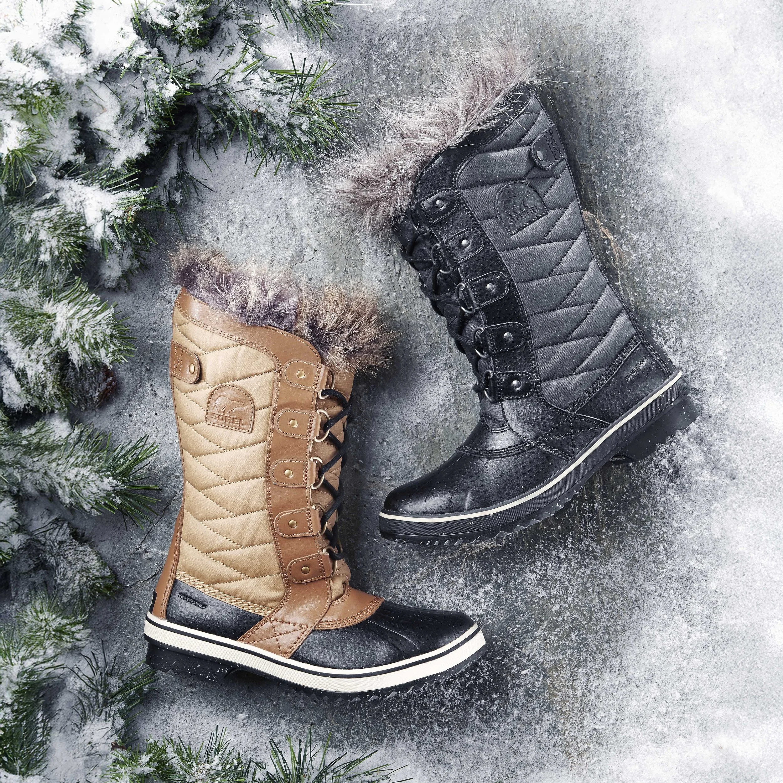 Two winter boots, one black and one tan, with fur trim, laid on snow-covered ground near a snow-dusted pine branch.
