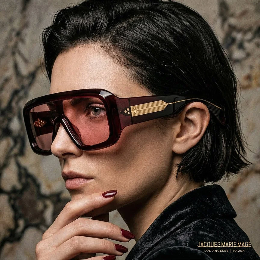 A woman with short black hair wearing futuristic sunglasses with pink tinted lenses and a black outfit, standing against a stone wall.