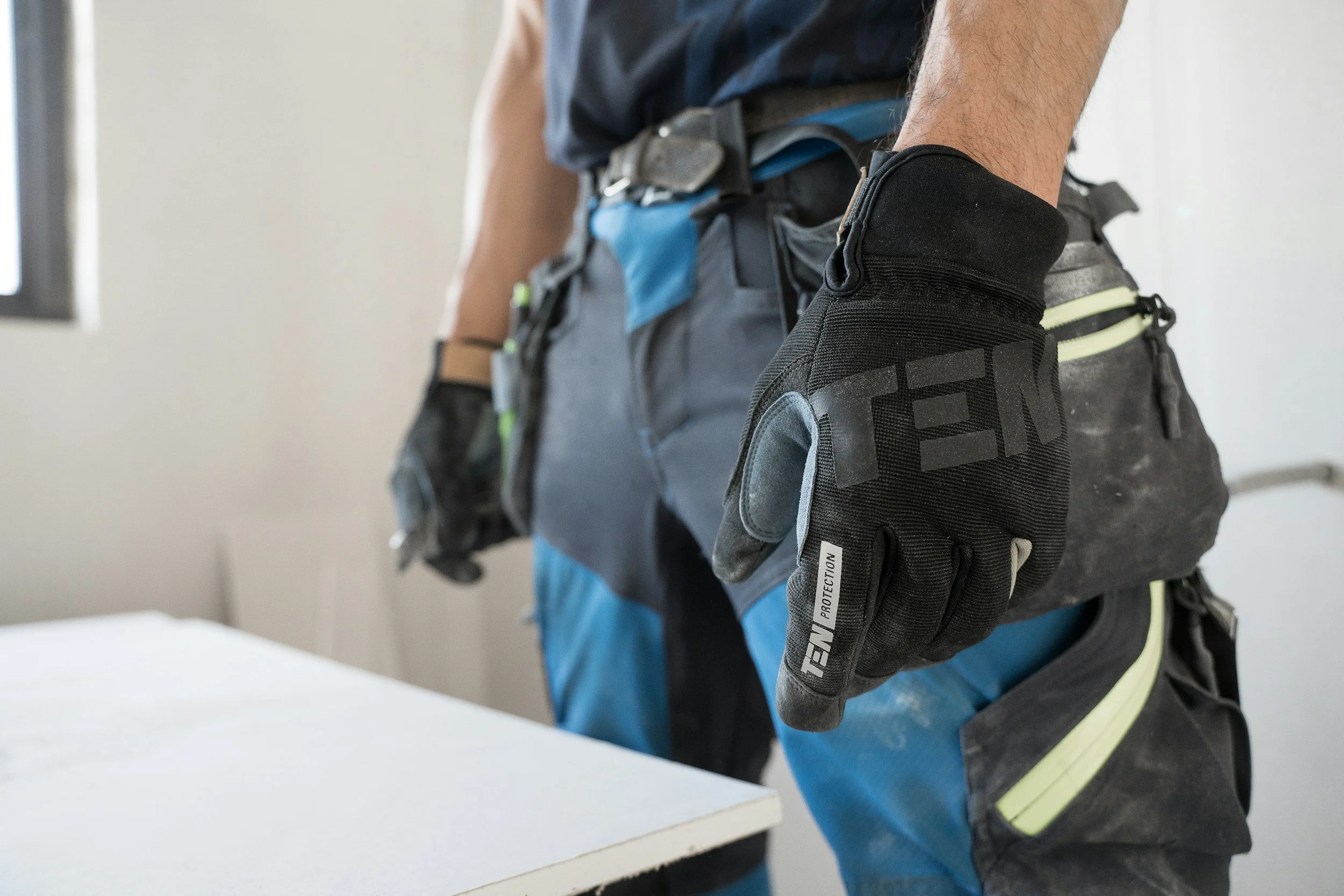 Close-up of a construction worker's gloved hand resting on a work surface, with safety gear and tool belt visible.