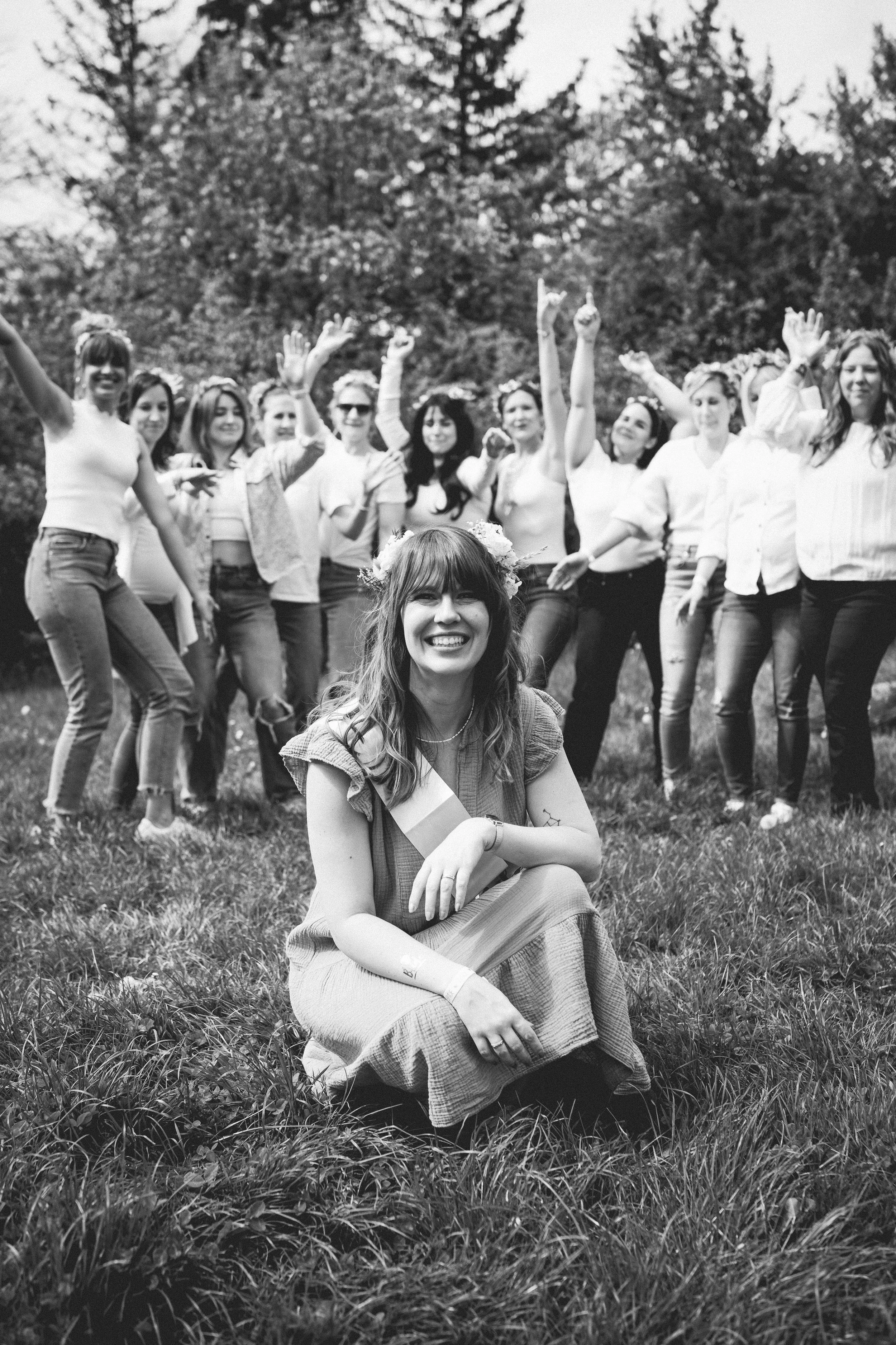 Eine Gruppe junger Frauen, die im Freien stehen und feiern, eine Frau sitzt vorne mit einer Blumenkette im Haar und trägt eine Sash, lachend.