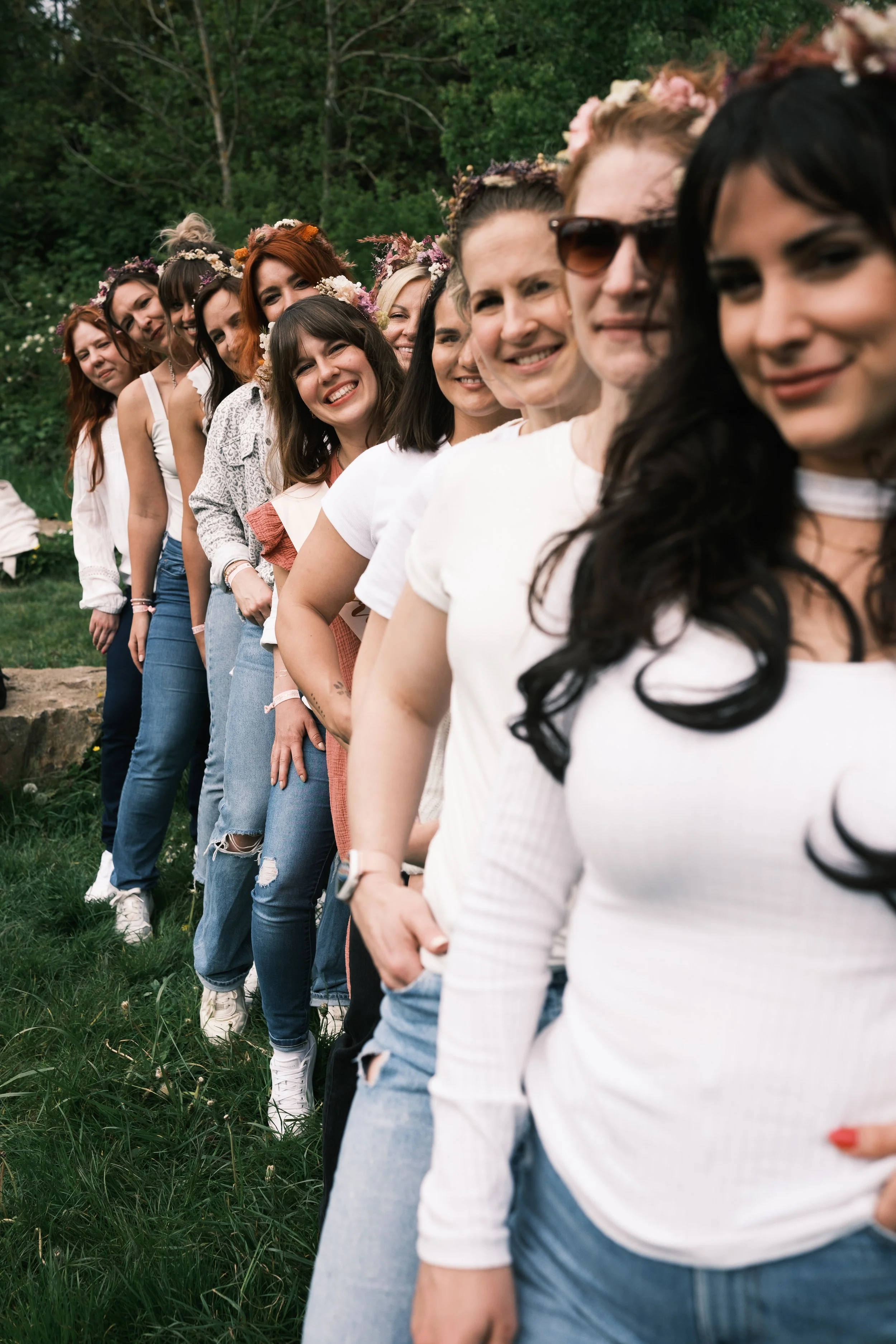 Eine Gruppe von Frauen steht in einer Schlange im Freien, viele tragen Blumenkronen, und alle sind lächelnd.