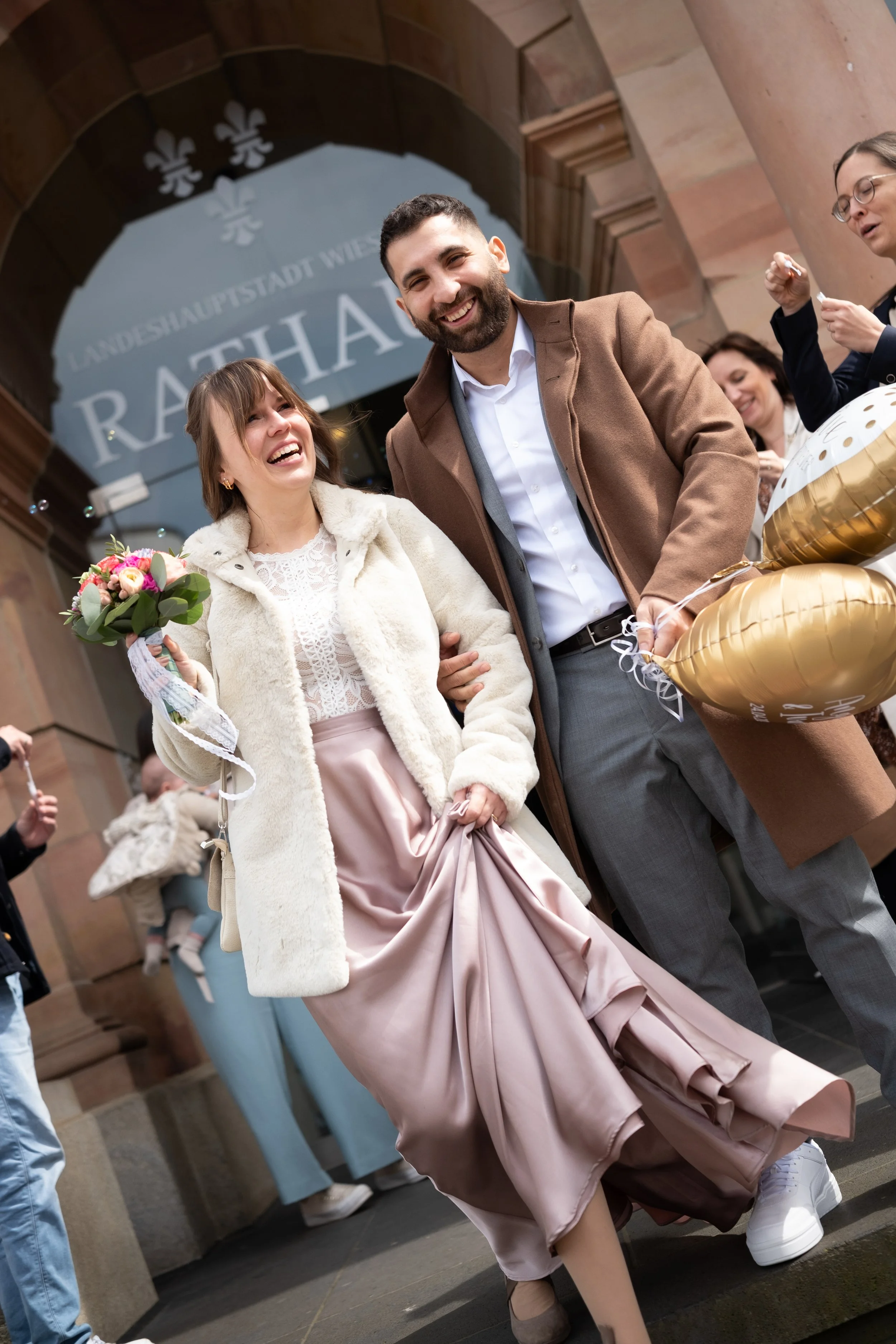Ein glückliches Paar bei einer Feier vor einem Rathaus, die Frau trägt ein langes rosa Kleid und hält einen Blumenstrauß, der Mann trägt einen Mantel und lächelt.