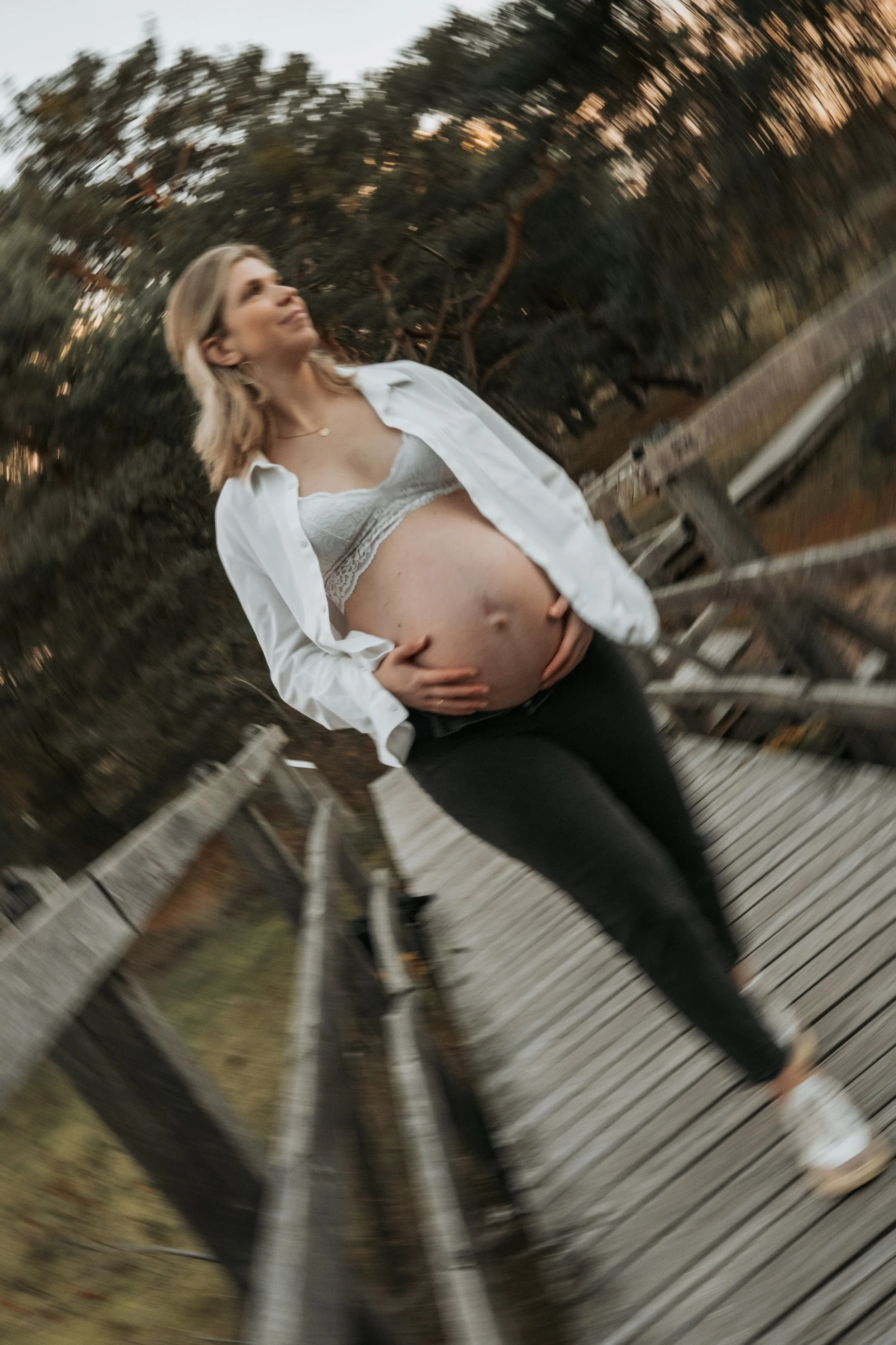 Schwangere Frau steht auf einer hölzernen Brücke im Freien mit Bäumen im Hintergrund, trägt off-shoulder weiße Bluse und schwarze Hose, lächelt und hält ihren Bauch.