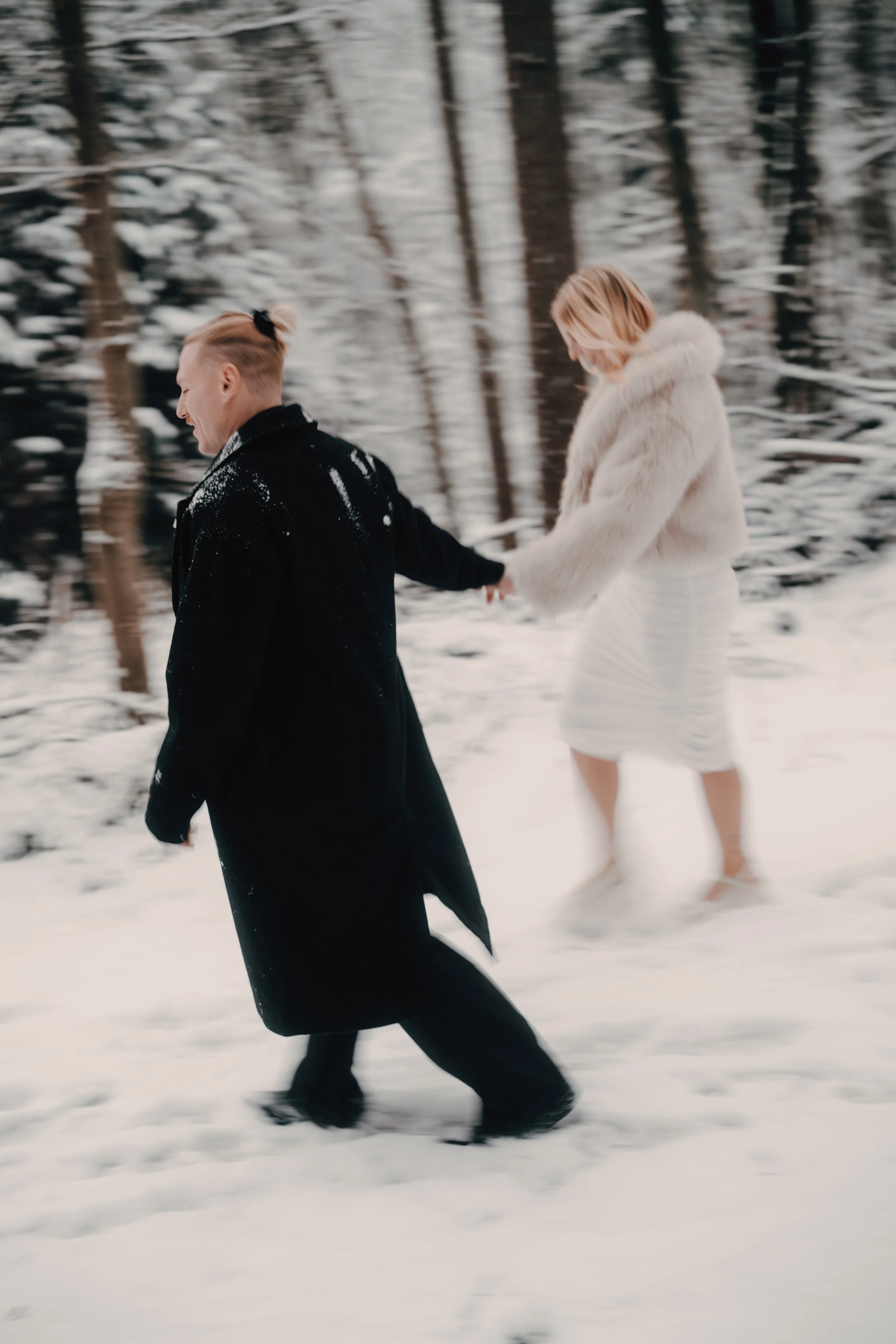 Ein Paar hält sich an einer Schneelandschaft im Wald an den Händen, die Frau trägt eine weiße Kleid und einen Pelzmantel, während der Mann einen schwarzen Mantel trägt.