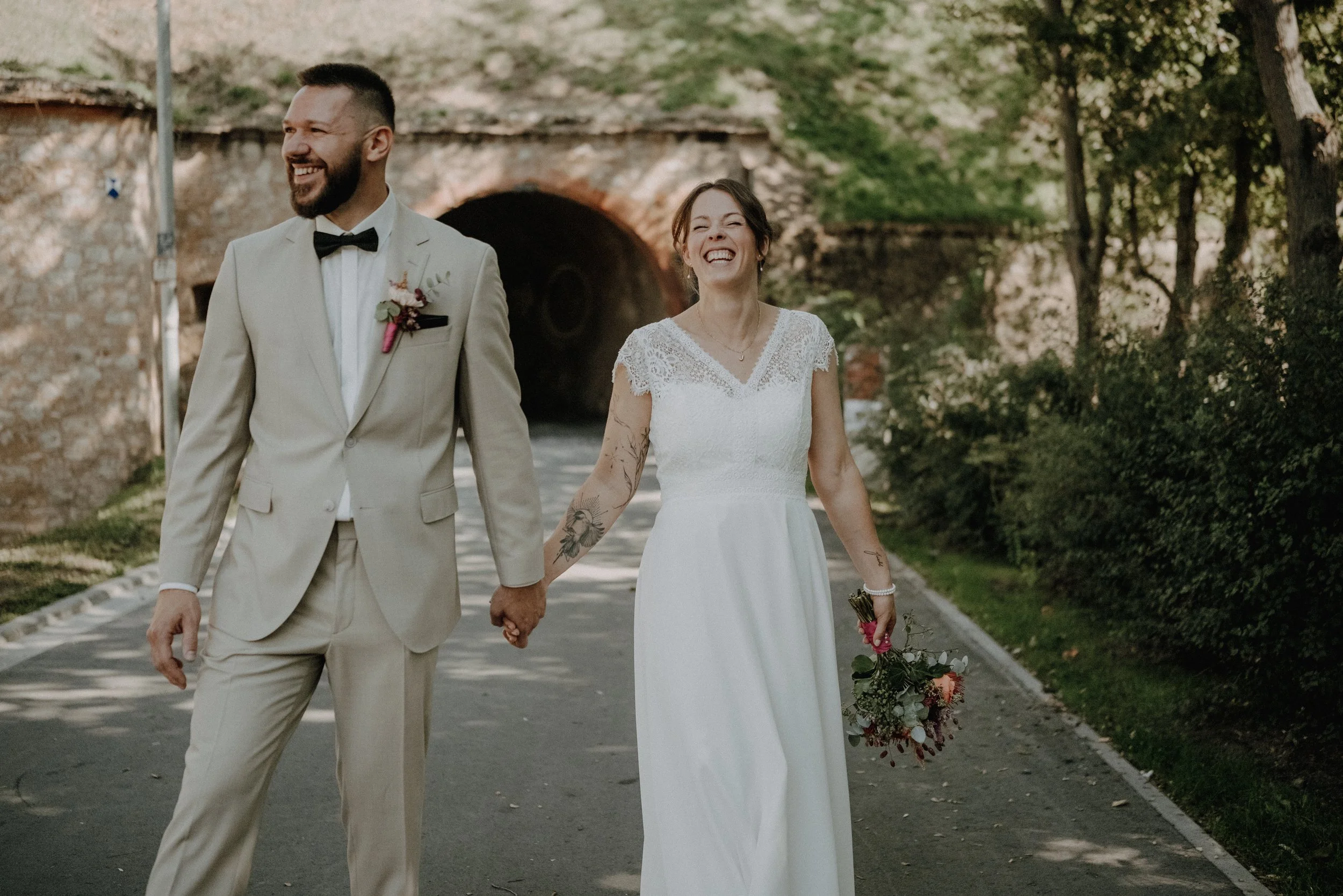 Ein lachendes Brautpaar in Hochzeitskleidung läuft in einer parkähnlichen Umgebung. Der Bräutigam trägt einen cremefarbenen Anzug mit Fliege, die Braut trägt ein weißes Hochzeitskleid mit Spitzenverzierungen und hält einen Blumenstrauß. Beide halten 