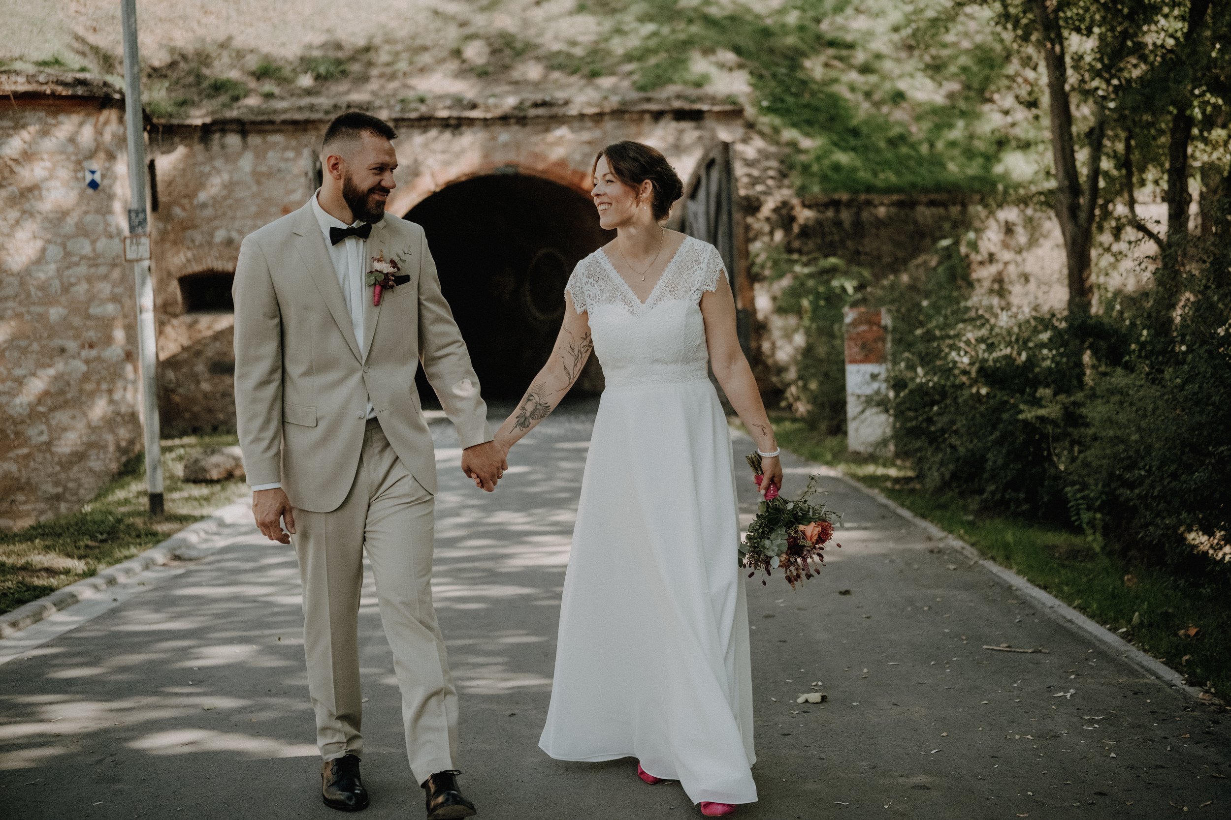 Ein frisch verheiratetes Paar läuft Hand in Hand auf einer Straße, mit einem Tunnel im Hintergrund. Sie lächeln sich liebevoll an, die Frau trägt ein weißes Hochzeitskleid und hält einen Blumenstrauß, der Mann trägt einen beige Anzug mit Fliege.