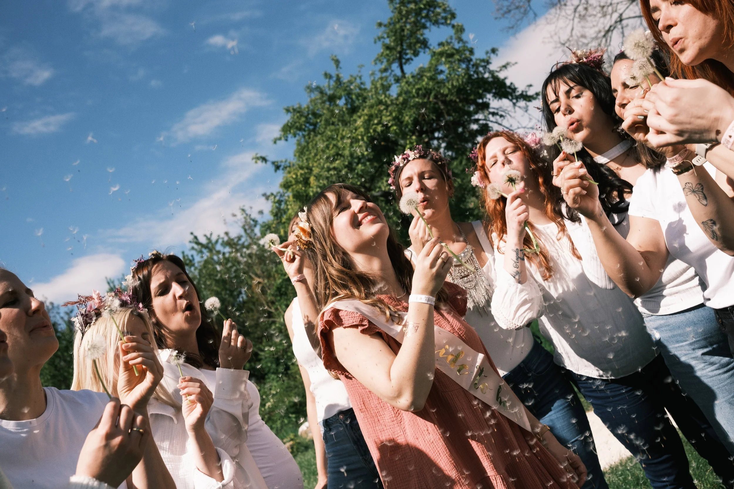 Eine Gruppe von Frauen im Freien, die Dandelion-Samen pusten, um einen Abschluss oder eine Feier zu feiern, mit blauen Himmel und grünen Bäumen im Hintergrund.