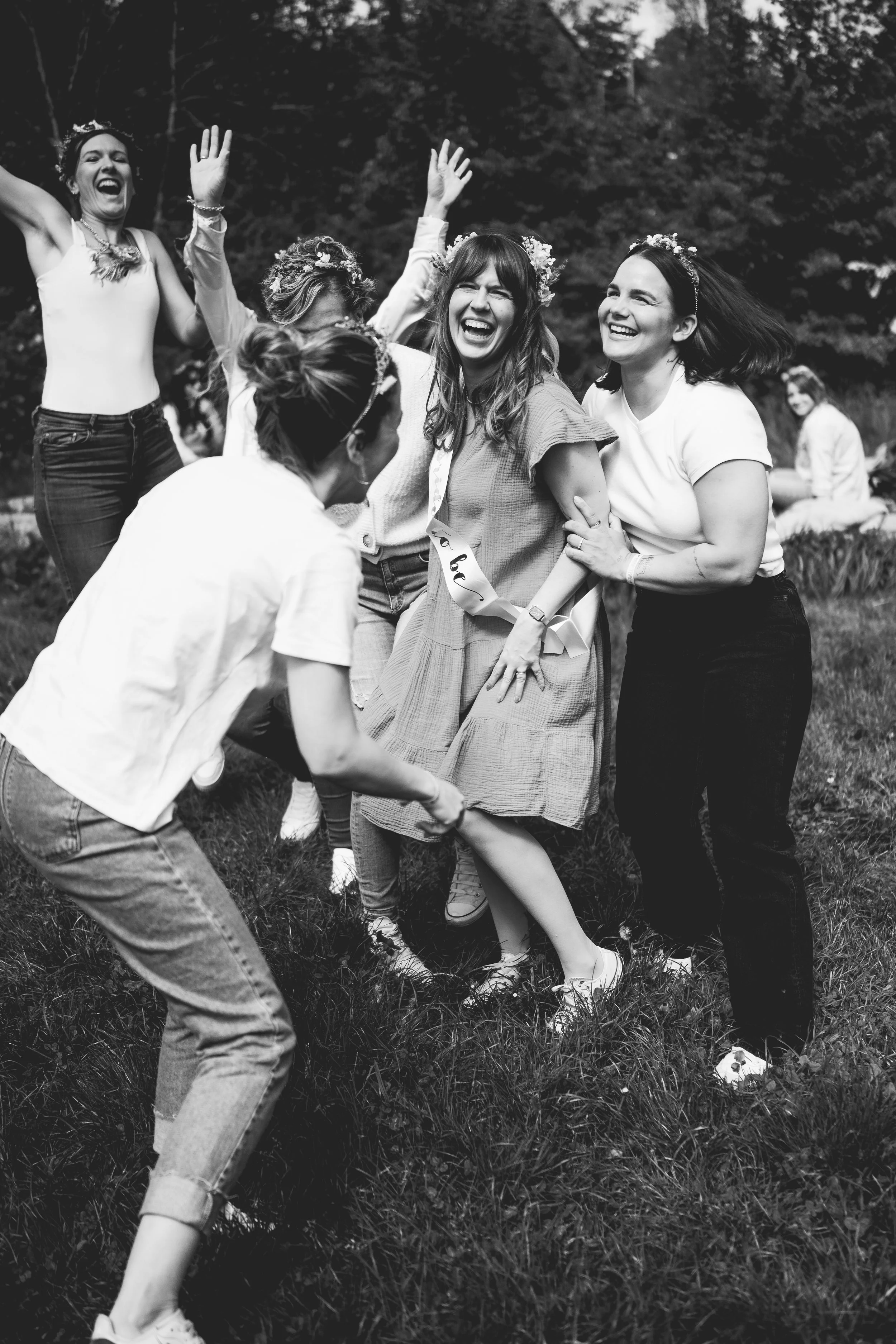 Gruppe fröhlicher Frauen auf einem Rasen im Freien, beim Feiern, mit lachenden Gesichtern, einige tragen Blumenkränze im Haar, eine trägt ein 