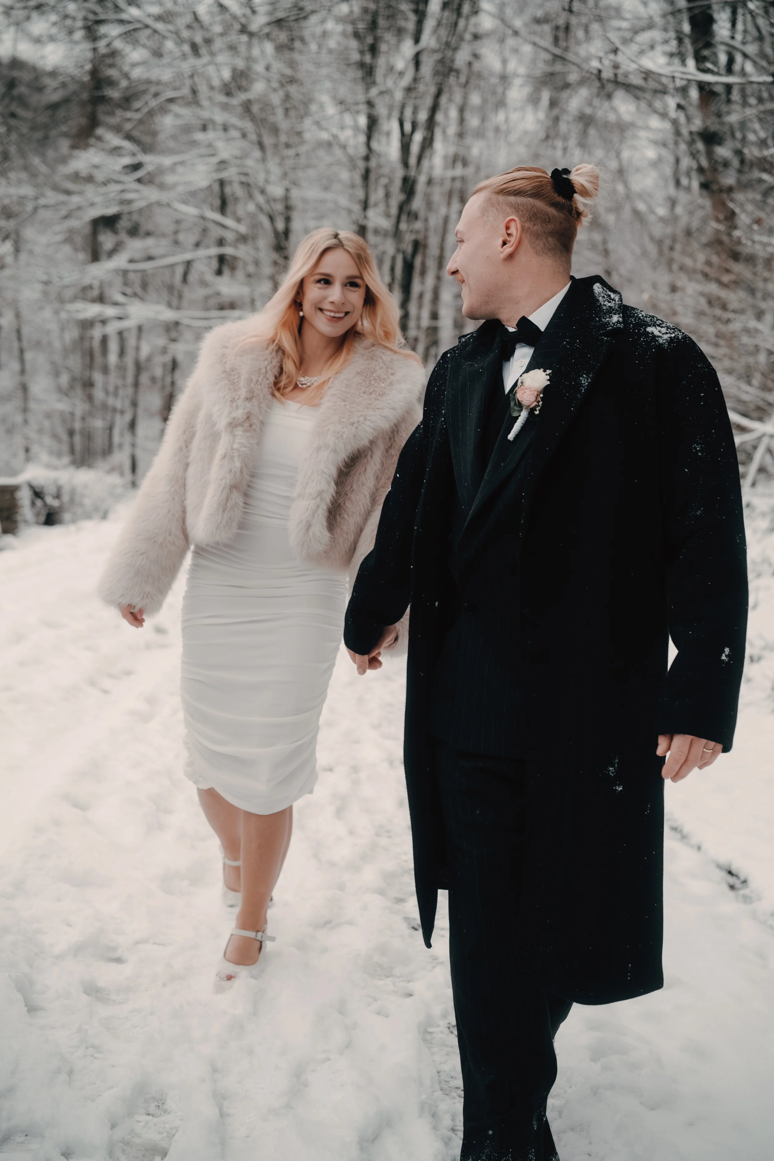 Ein heiratendes Paar spaziert im Schnee in einem winterlichen Wald, die Frau trägt ein weißes Kleid und Pelzjacke, der Mann trägt einen schwarzen Anzug mit Mantel und Fliege.