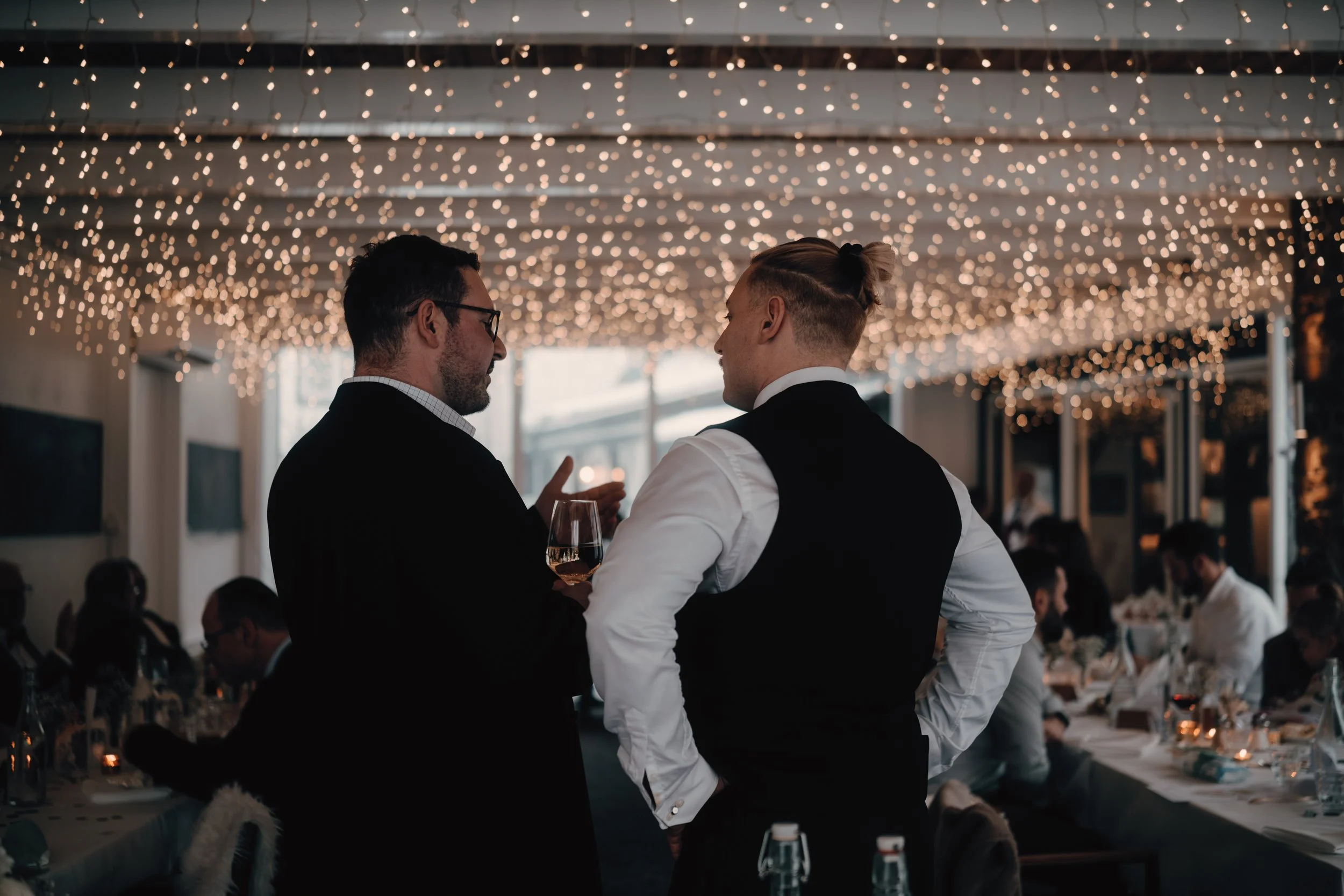Zwei Männer beim Gespräch auf einer festlichen Feier, umgeben von Tischgästen, mit Decken aus Lichterketten an der Decke im Hintergrund.