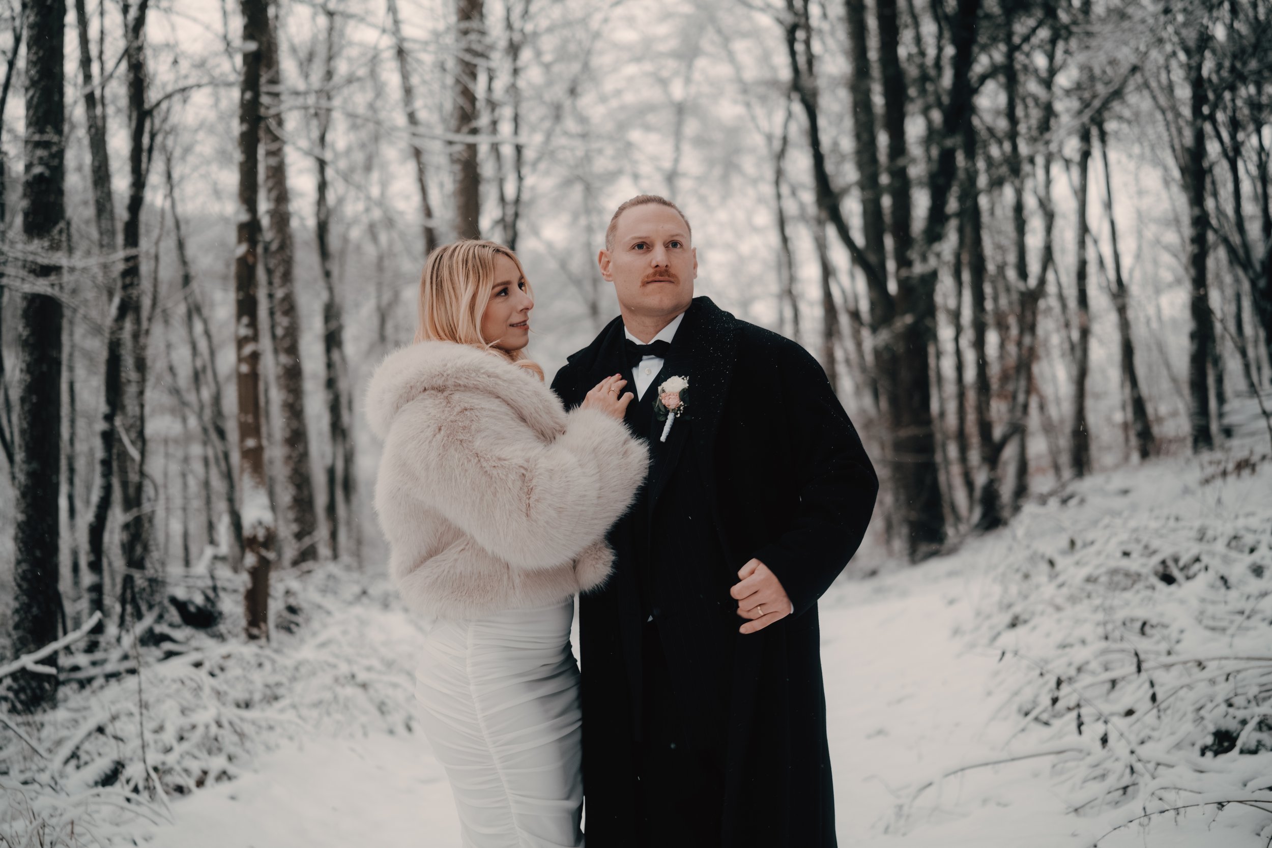 Ein Paar in formeller Kleidung steht in einem verschneiten Wald, vermutlich anlässlich einer Hochzeit.