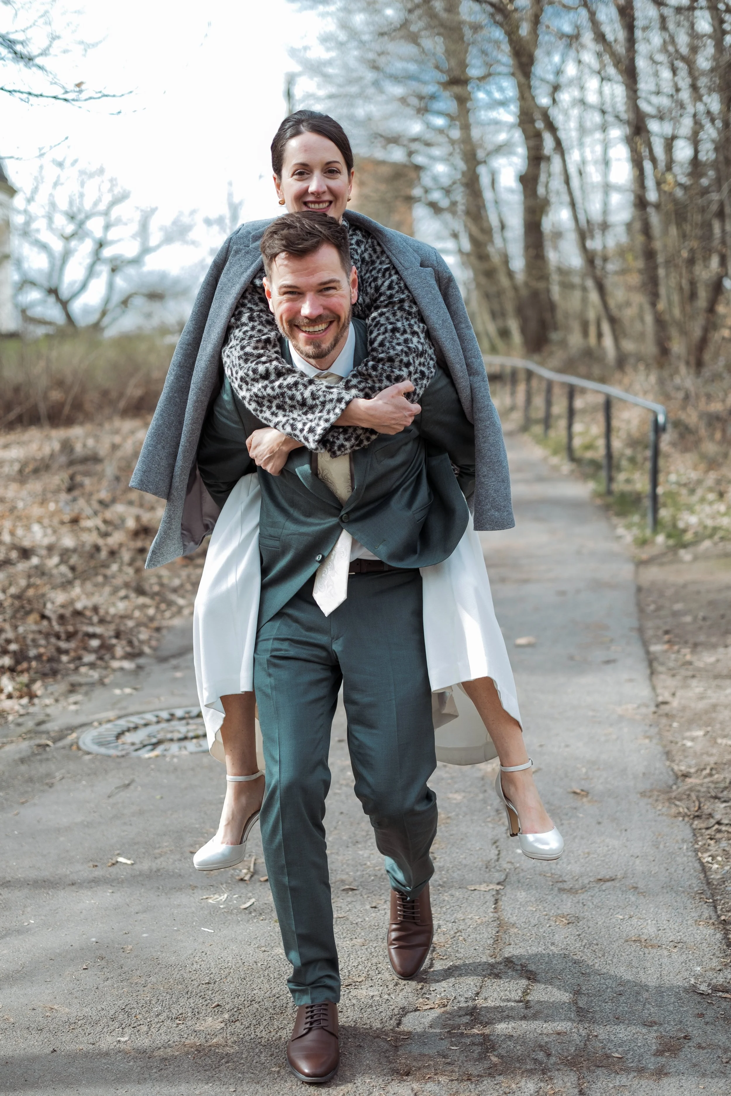 Ein Mann trägt eine Frau auf den Schultern, beide sind glücklich und lachen, während sie auf einem Weg im Freien unterwegs sind.