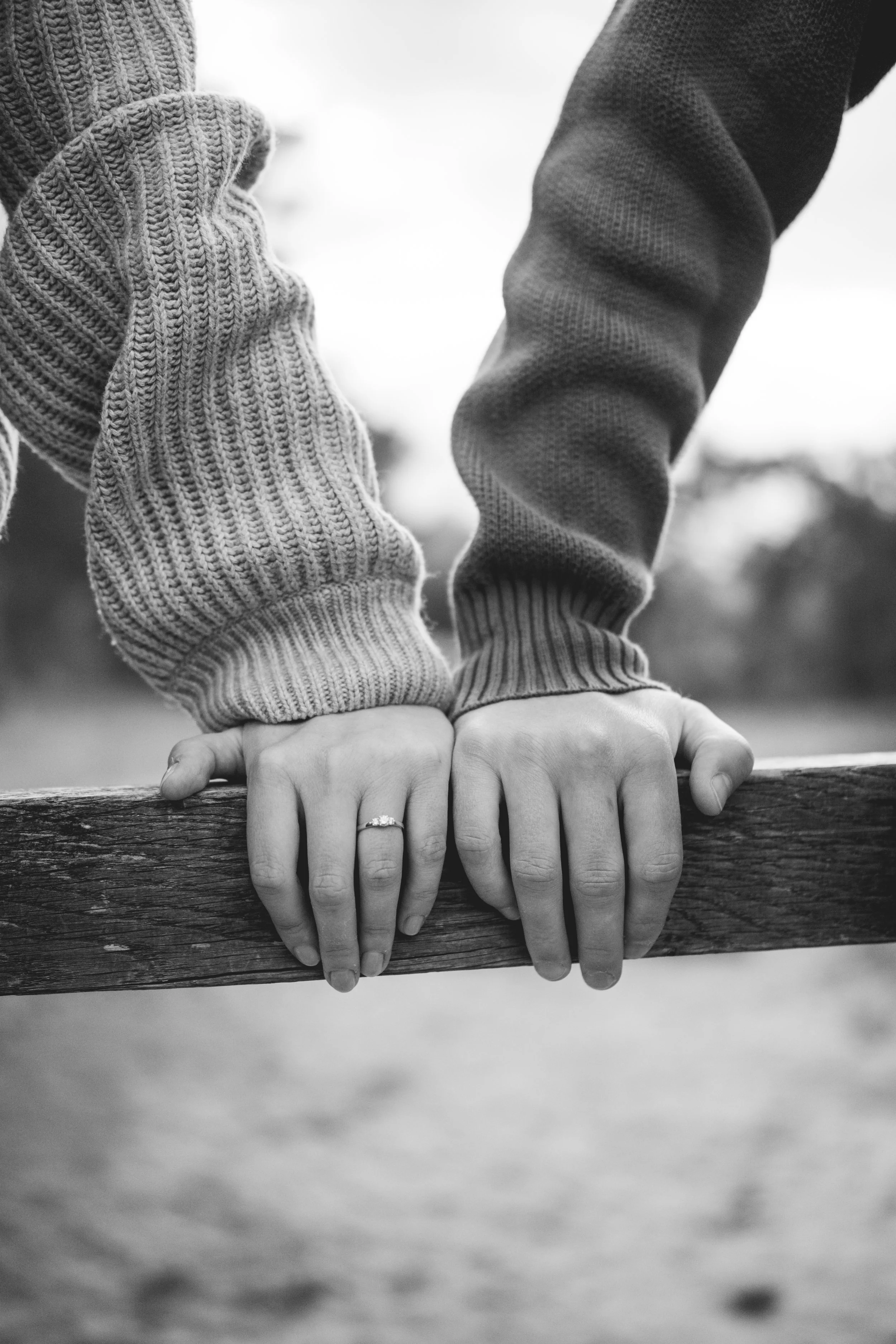 Zwei Hände mit Ringen, die auf einer Holzstange liegen, in schwarz-weiß. Eine Person trägt einen Strickpullover, die andere einen Pullover mit kurzen Ärmeln.