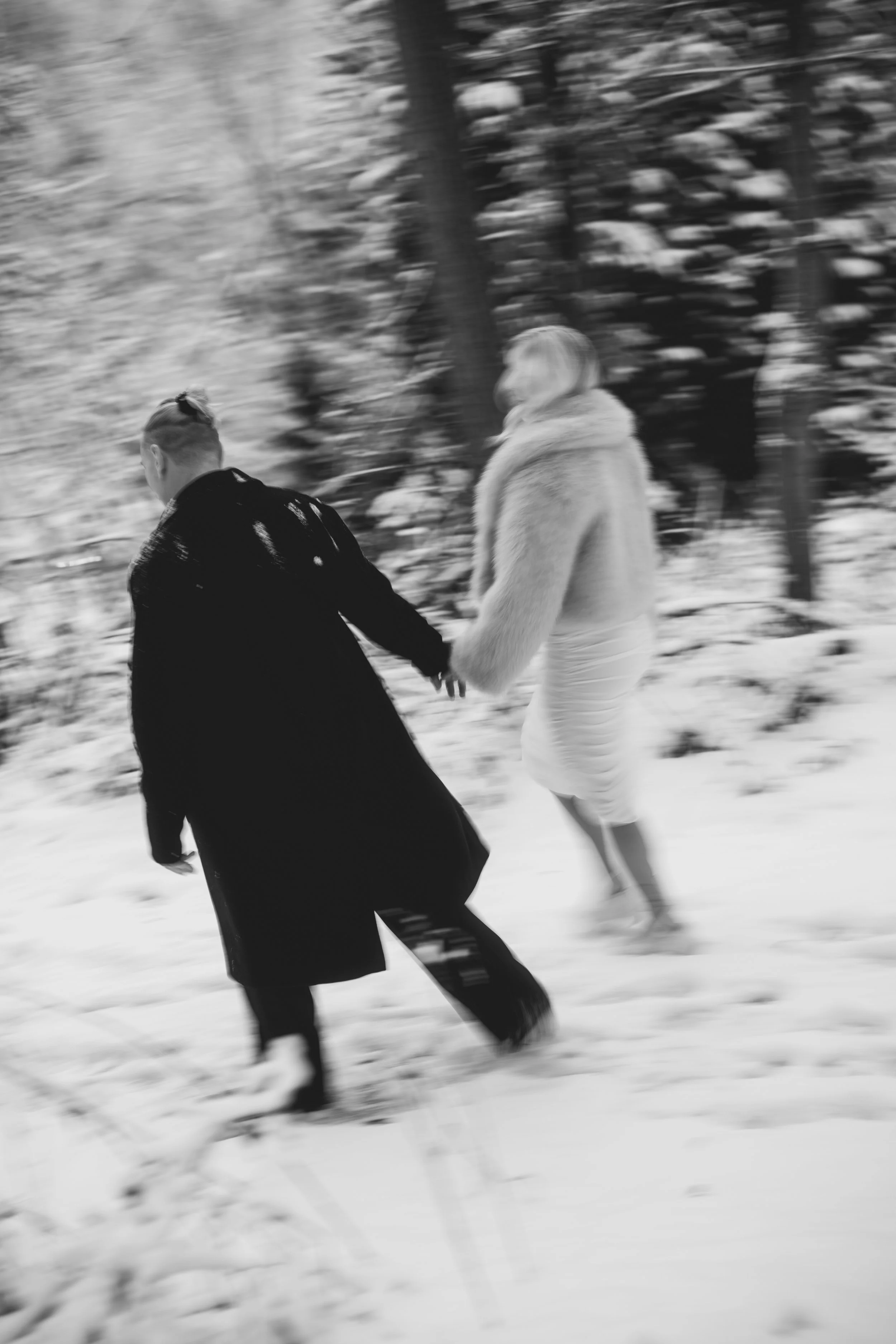 Zwei Frauen, eine in einem schwarzen Mantel und die andere in einem weißen Kleid mit Felljacke, laufen durch den Schnee im Wald, Hand in Hand.