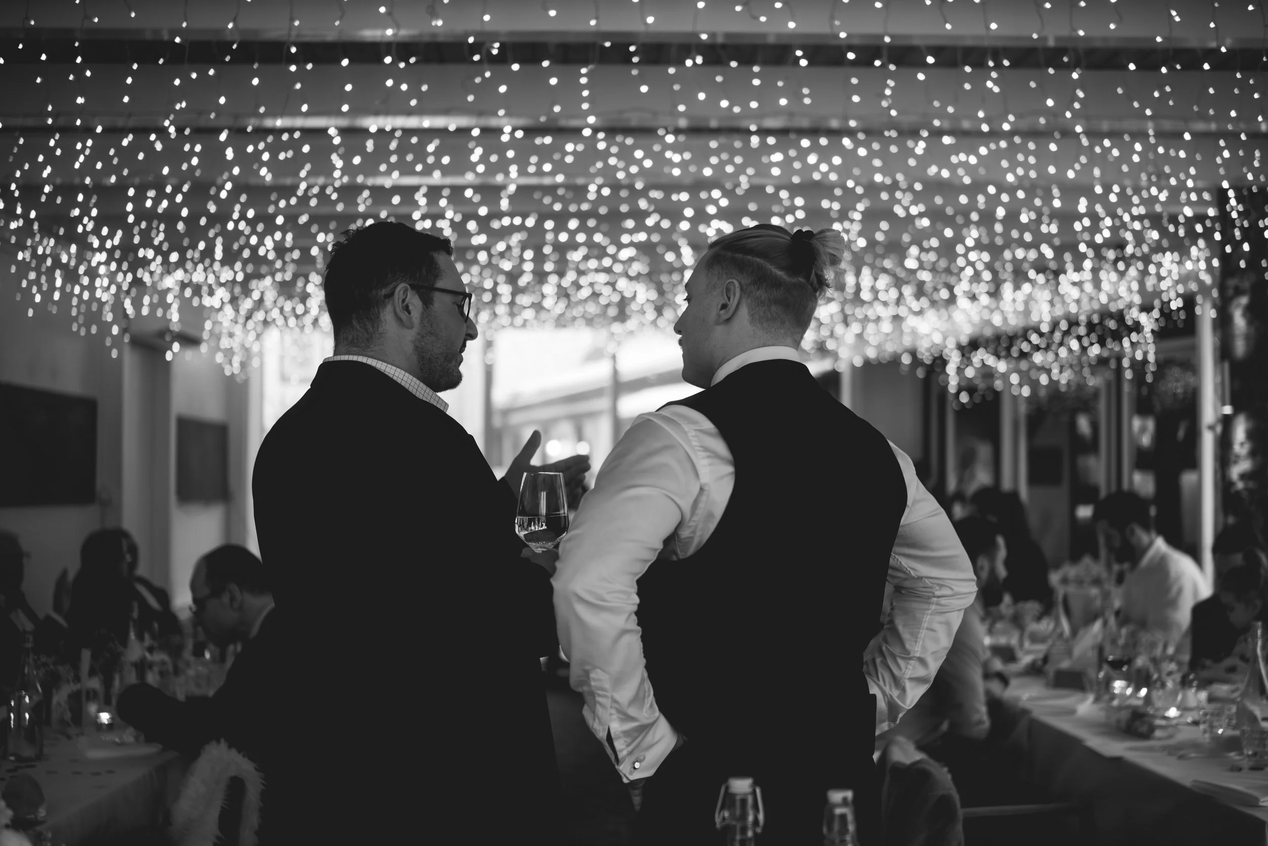 Zwei Männer im Gespräch bei einer festlichen Abendveranstaltung, beleuchtet von festlichen Lichterketten.