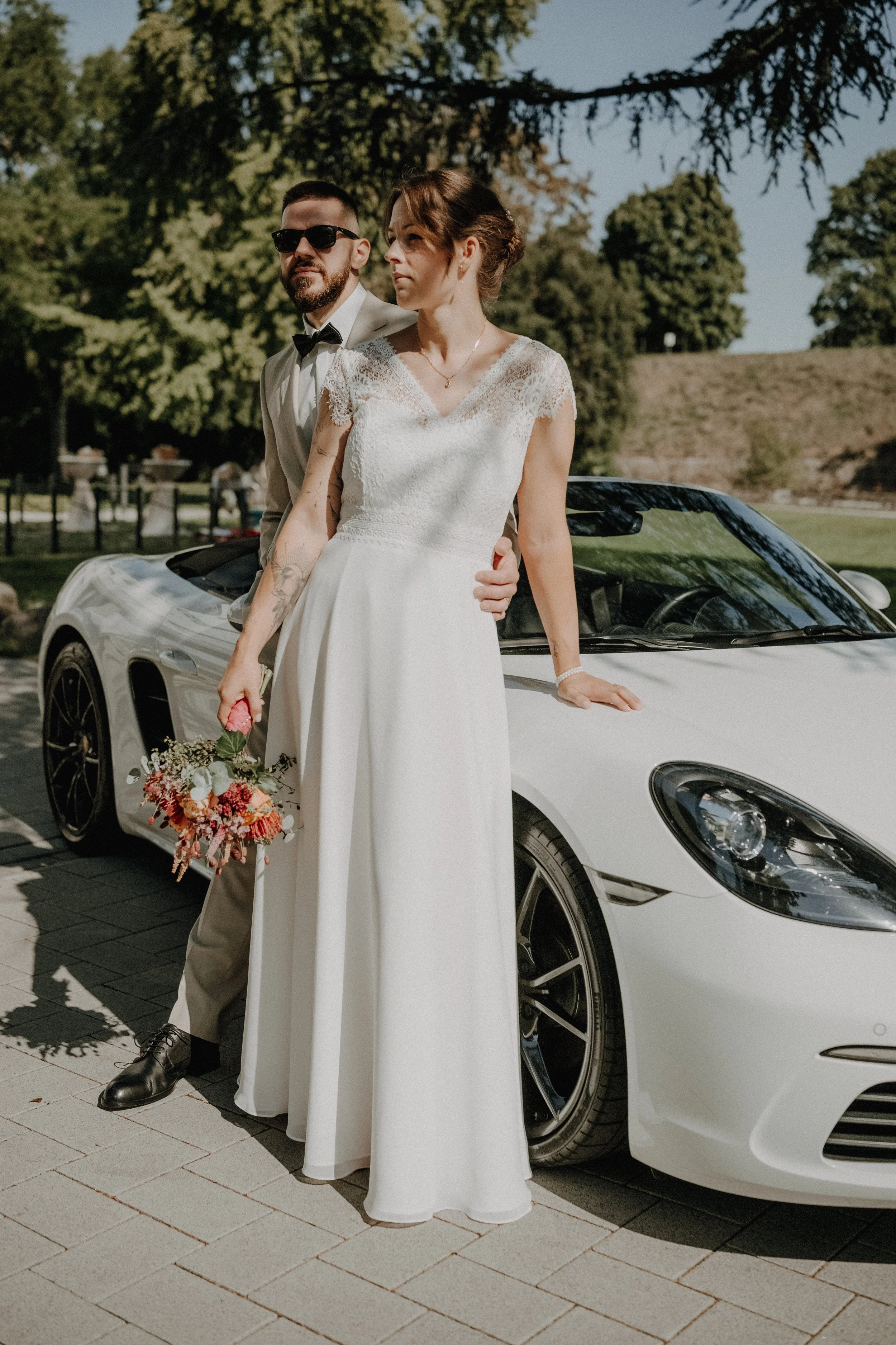 Ein Brautpaar steht neben einem weißen Sportwagen im Freien, die Frau hält einen Blumenstrauß, der Mann trägt einen Anzug mit Fliege, es ist sonnig, grüne Bäume im Hintergrund.