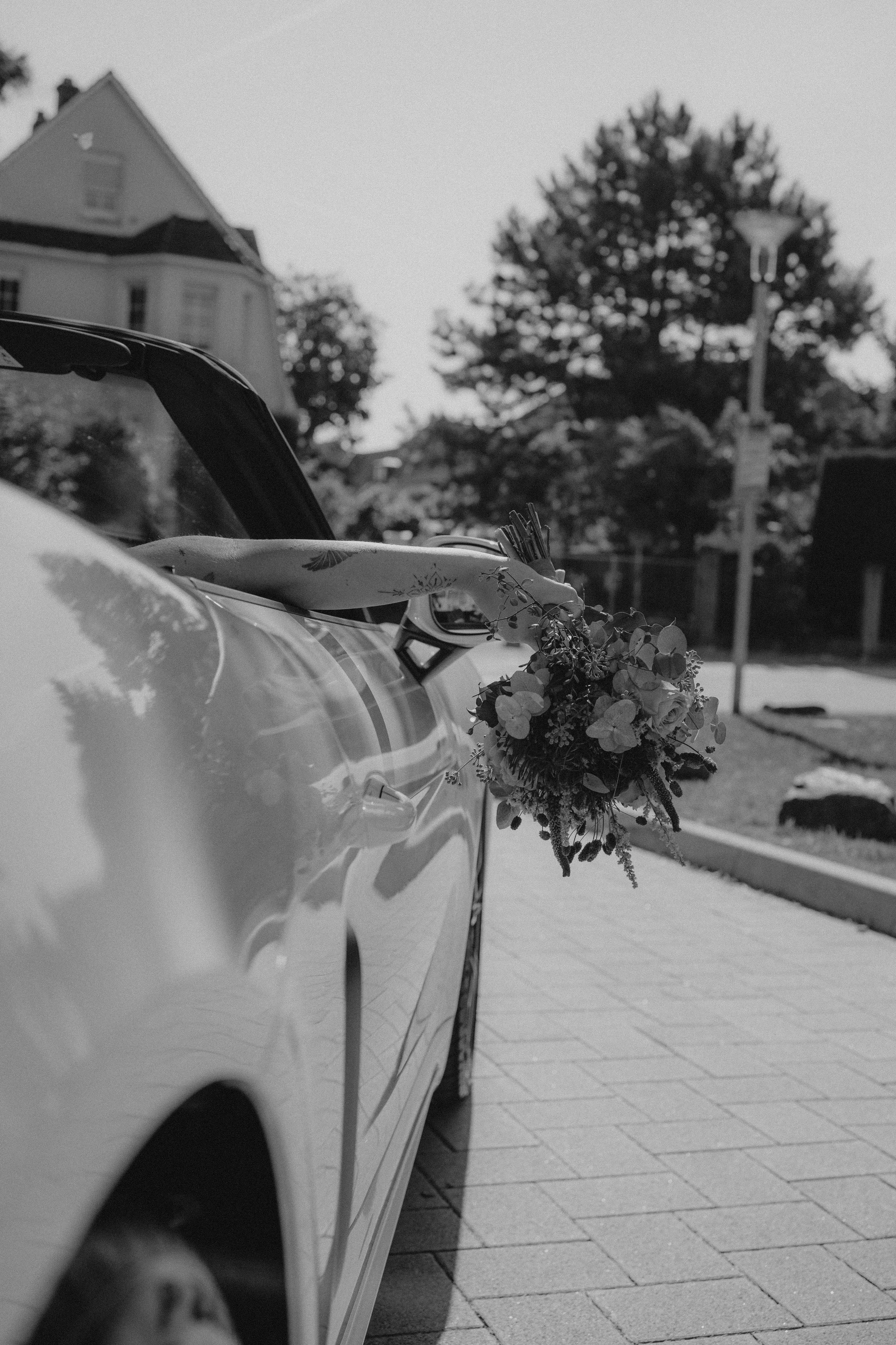 Ein Auto, an dem ein Blumenstrauß aus dem offenen Kofferraum hängt, auf einer gepflasterten Straße, im Hintergrund Bäume und ein Haus in Schwarz-Weiß.