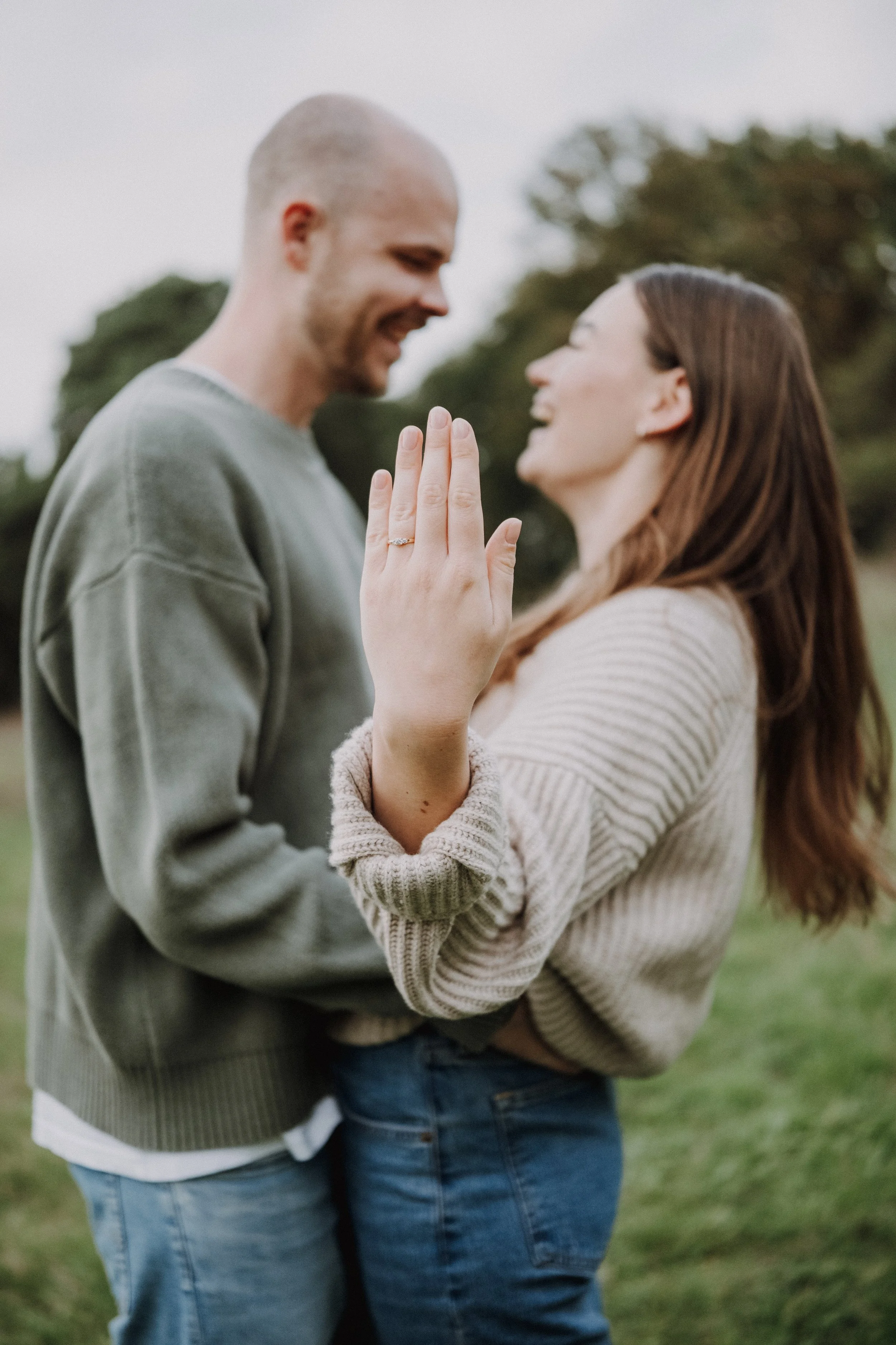 Ein Paar steht im Freien, lächelt sich an und zeigt ihre Hände mit einem Ehering, was auf eine Verlobung oder Heirat hinweist.