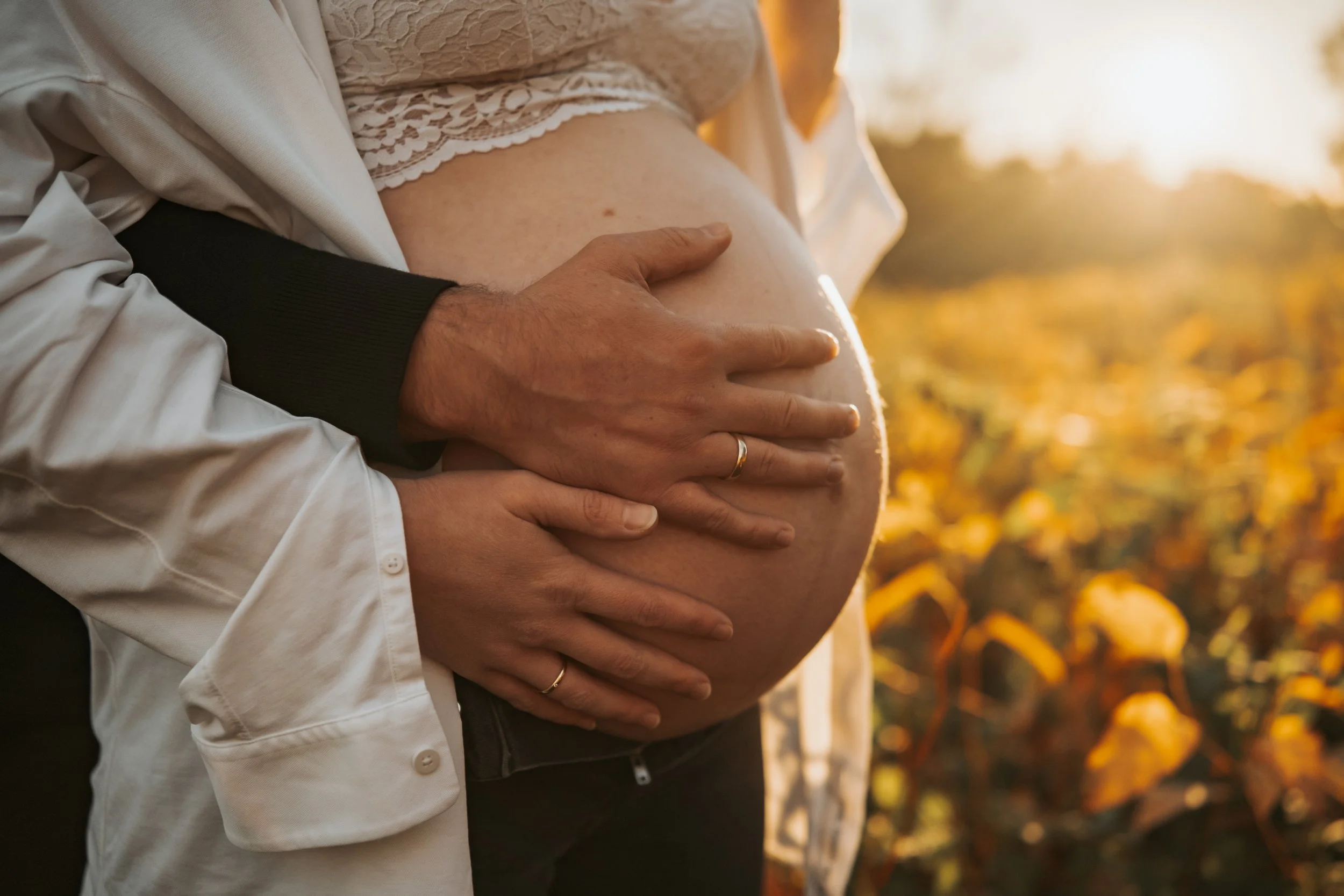 Ein schwangere Person wird von einer weiteren Person in einem Sonnenuntergang im Freien gestreichelt, beide tragen Ringe an den Händen.