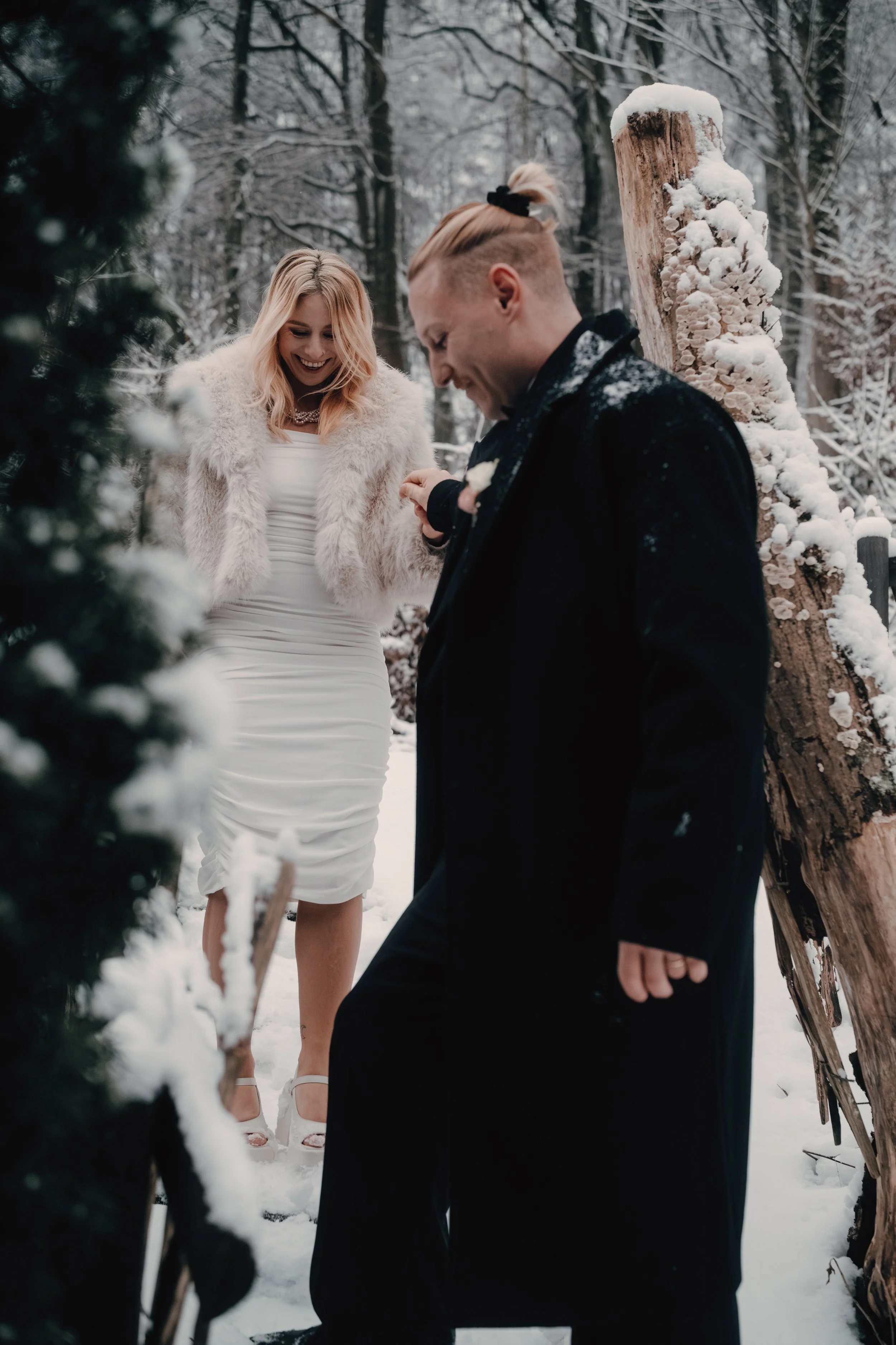 Ein Paar in formeller Kleidung steht im Schnee in einem verschneiten Wald, sie lachen und schauen auf etwas in ihren Händen, während der Mann einen schwarzen Anzug trägt und die Frau ein weißes Kleid mit Pelzjacke. Im Hintergrund ist ein geschmücktes