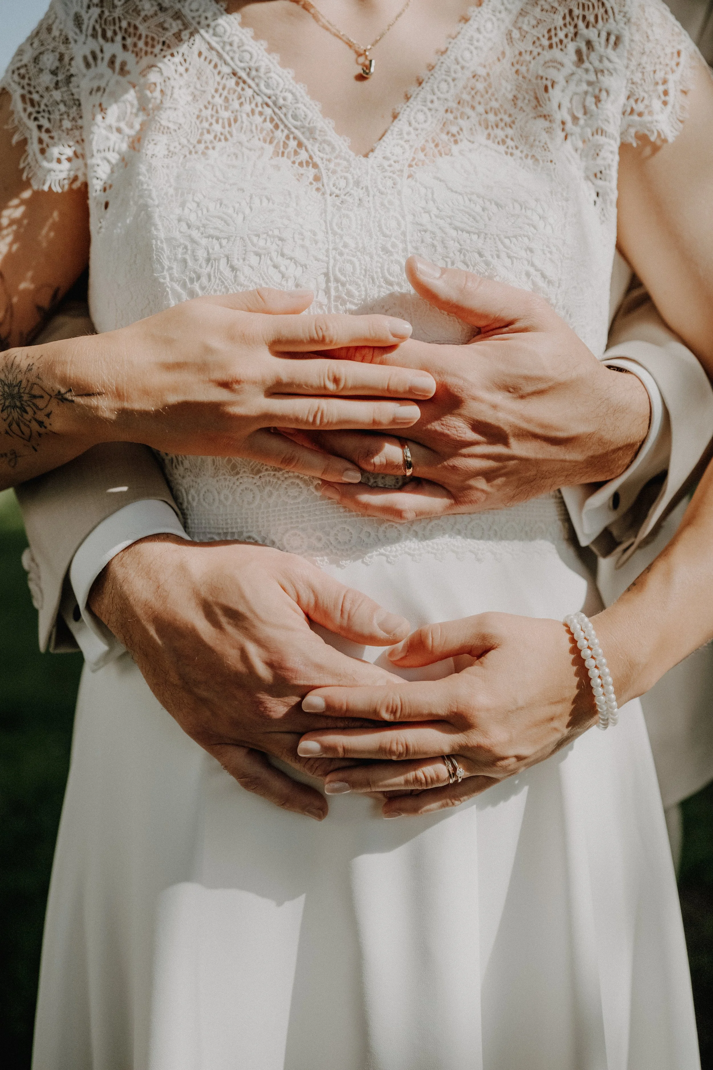 Nahaufnahme von vier Händen, die gemeinsam auf dem Bauch eines Menschen in einem weißen Kleid ruhen, mit Blick auf die Brautmutter, die an ihrer Hand eine Perlenarmband trägt, und die Braut, die einen Ehering trägt.
