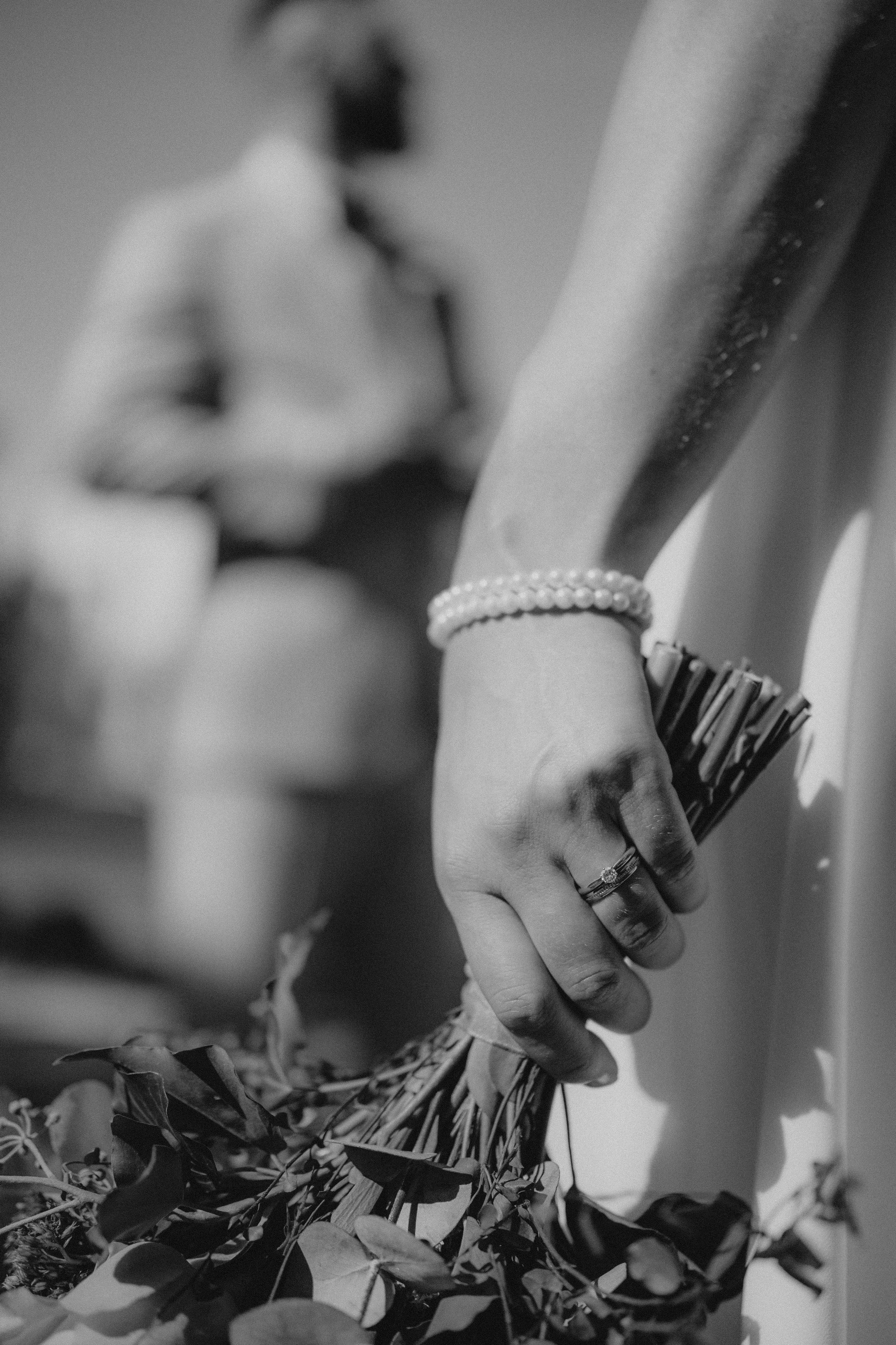 Nahaufnahme einer Hand mit Ring, Perlenarmband und Kette, die einen Strauß Blumen hält. Die Person im Hintergrund ist unscharf.