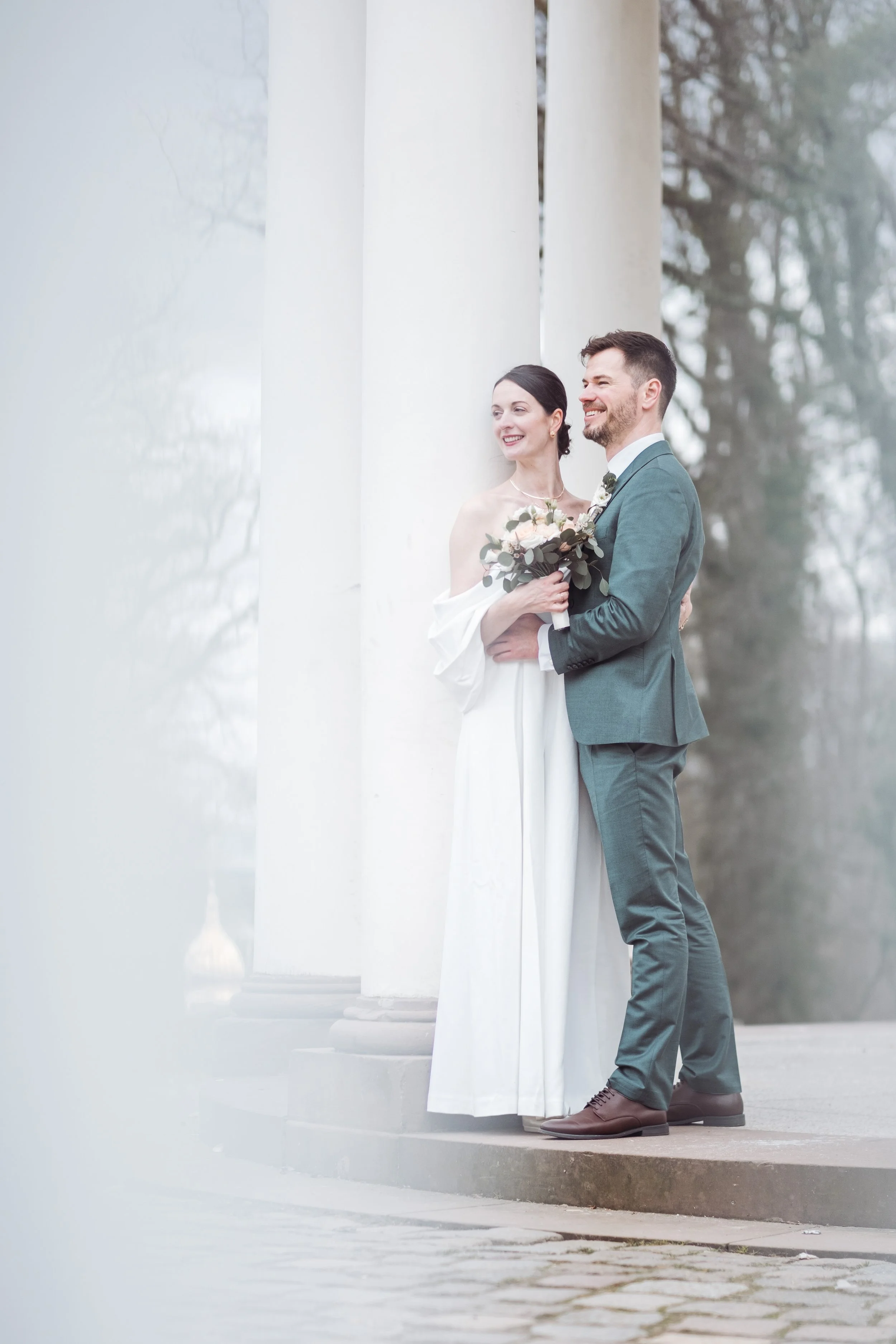 Ein glückliches Brautpaar steht vor weißen Säulen, die im Freien stehen. Die Braut trägt ein langes weißes Kleid und hält einen Blumenstrauß, während der Bräutigam in einem grauen Anzug steht.