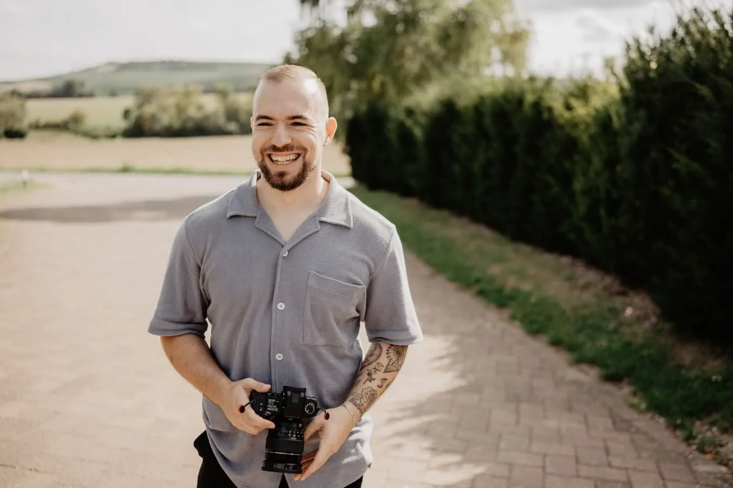 Ein lachender Mann mit Kamera in der Hand steht auf einem Weg im Freien, im Hintergrund sind Bäume und Hügel sichtbar.