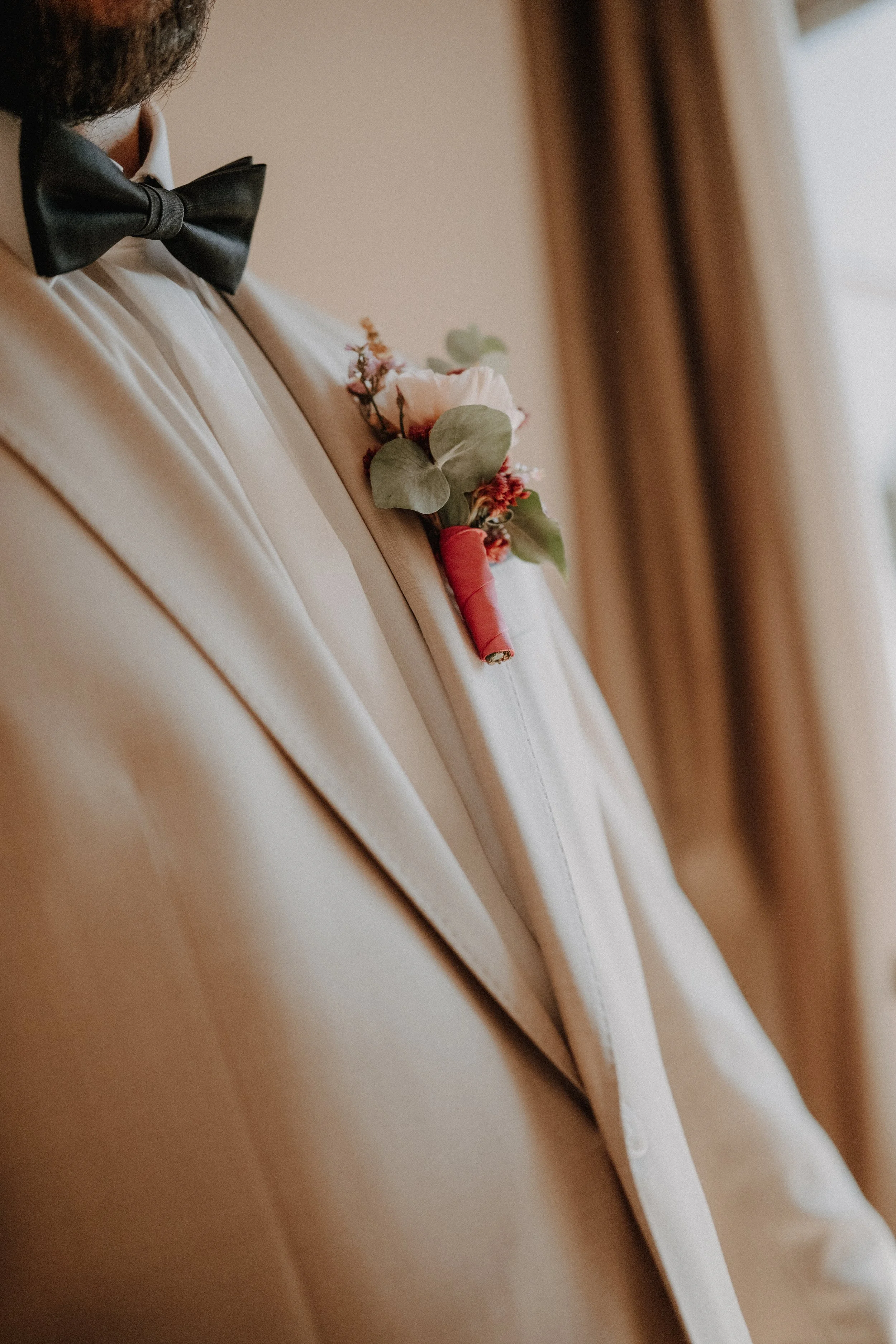 Ein Mann in weißem Anzug mit schwarzer Fliege trägt eine Boutonnière am Revers. Die Boutonnière besteht aus rosafarbenen Blüten, grünen Blättern und ist mit rotem Band umwickelt.