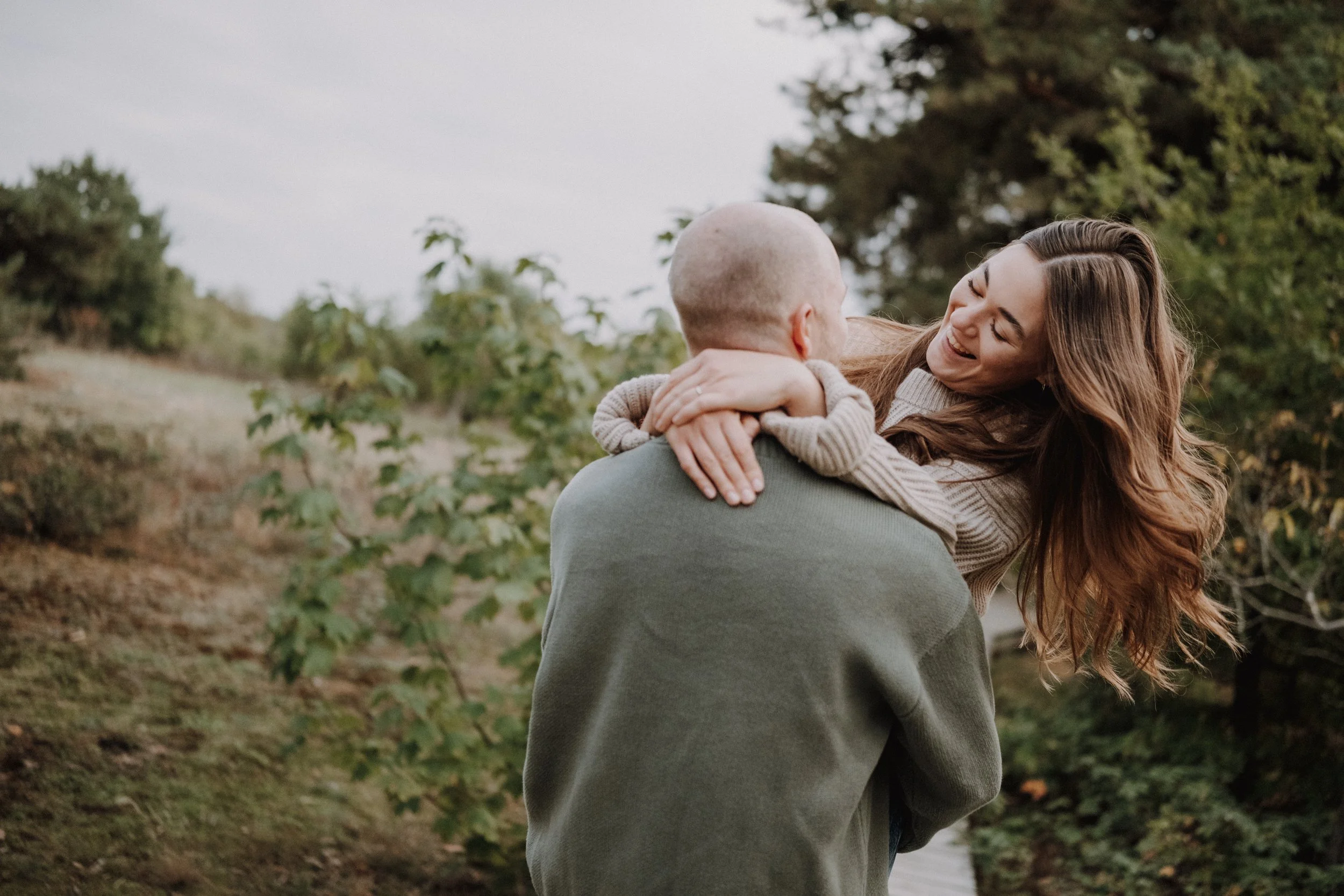 Ein Mann trägt eine lachende Frau, die ihn umarmt, im Freien mit Bäumen im Hintergrund.