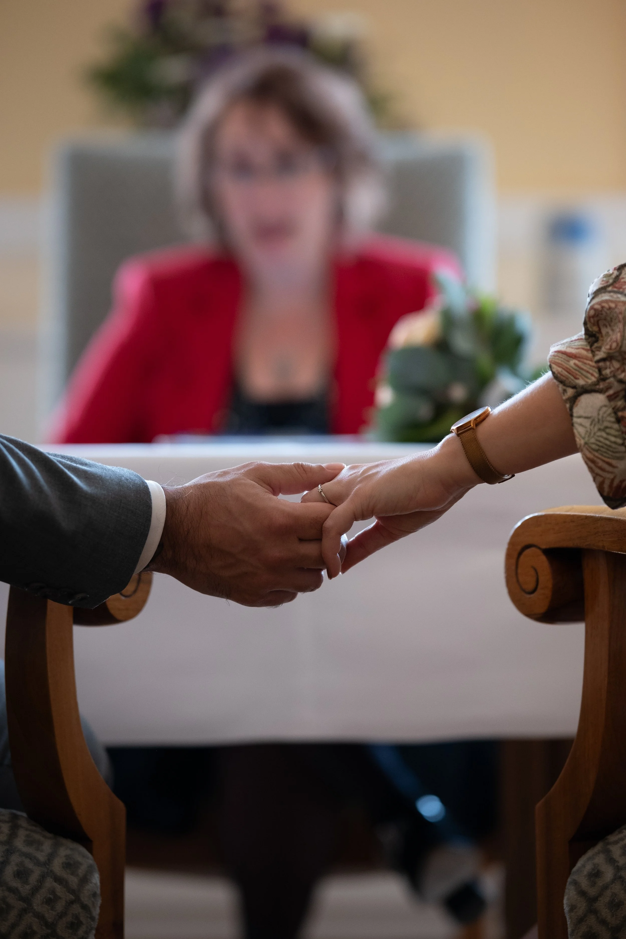 Ein Mann und eine Frau halten sich bei einer Zeremonie an den Händen, im Hintergrund eine Frau in einem roten Kleid, unscharf.