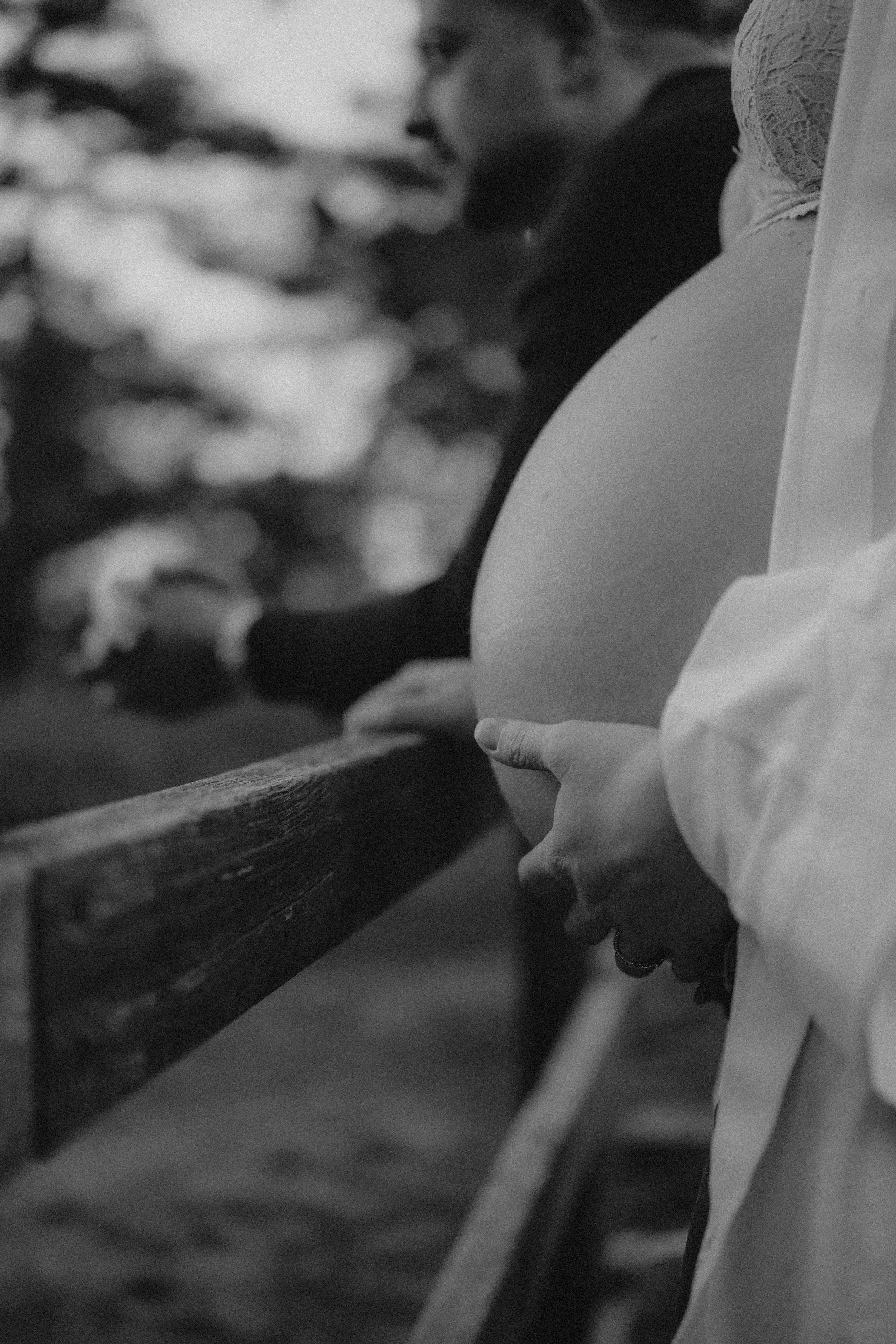 Nahaufnahme eines schwangeren Bauches, gehalten von einer Hand, mit einer Person im Hintergrund, die auf eine Wasseroberfläche blickt, in Schwarz-Weiß.