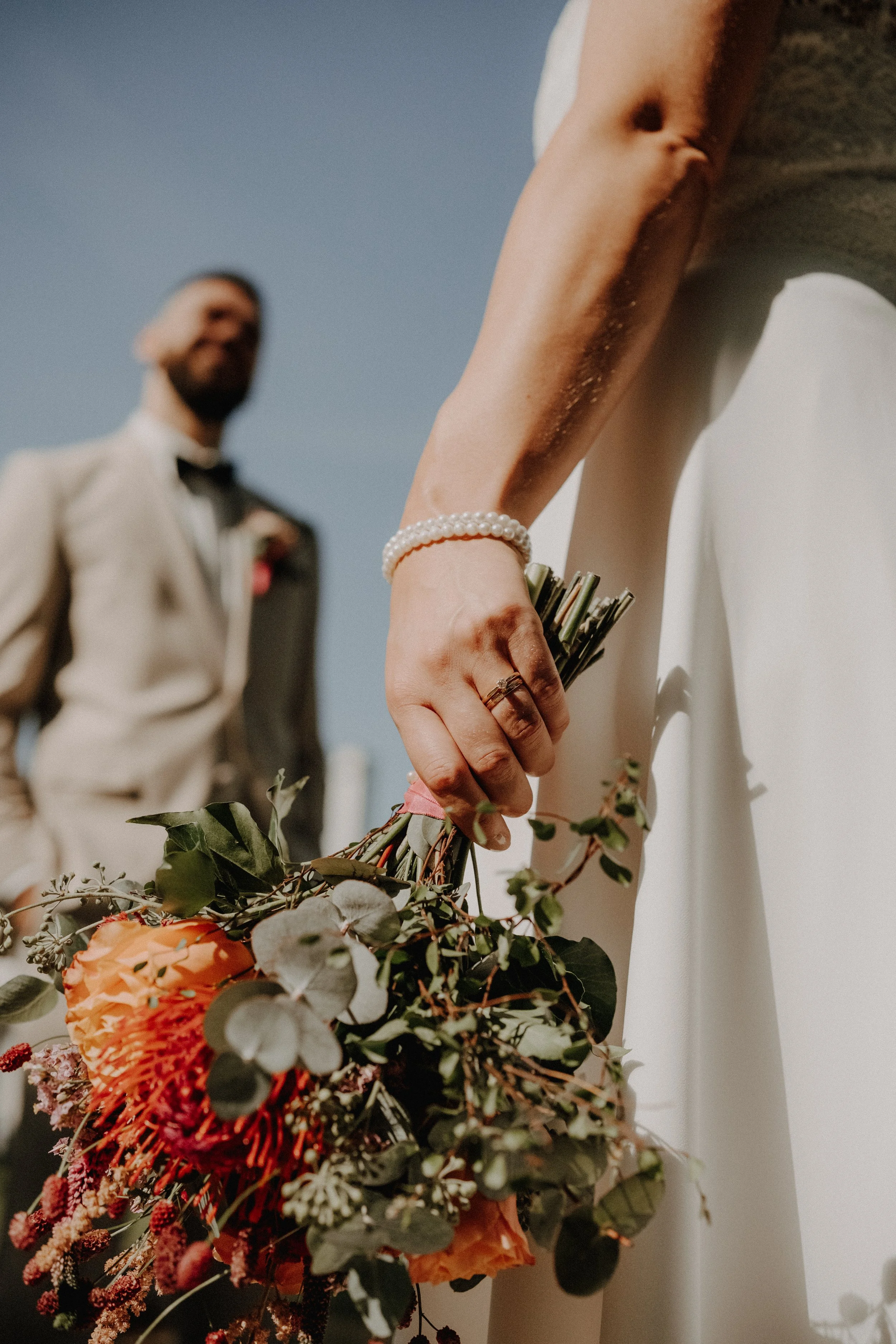 Fokus auf eine Hand einer Braut, die einen Brautstrauß hält, während im Hintergrund ein Bräutigam zu sehen ist, bei einer Hochzeit im Freien bei sonnigem Wetter.